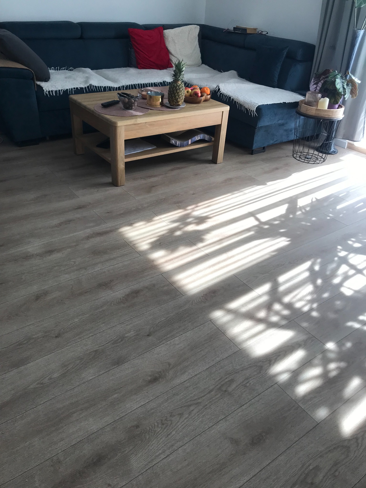 Ułożone panele podłogowe w salonie z widocznym fragmentem sofy, stolika kawowego i padającego światła słonecznego tworzącego cienie na podłodze.