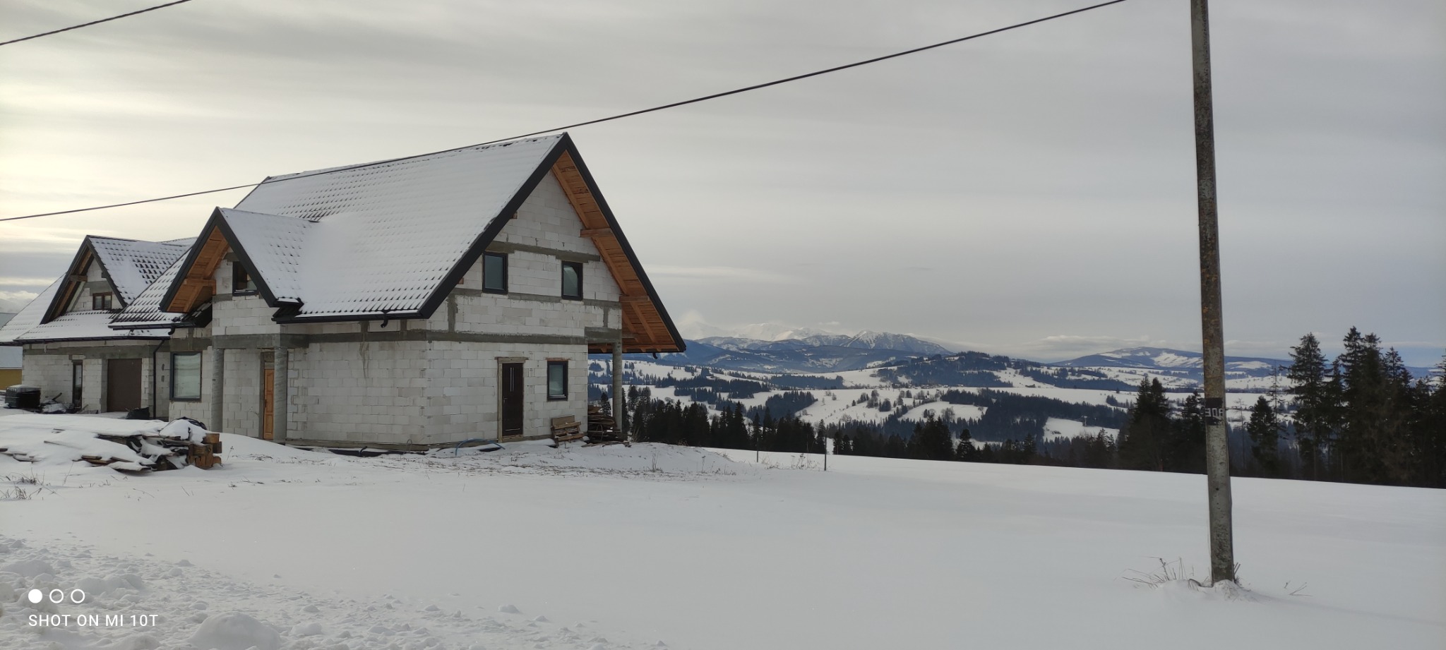 Surowy dom z białego bloczka w zimowym krajobrazie górskim, częściowo ośnieżony dach, widoczne drewniane elementy konstrukcji dachu, w tle ośnieżone szczyty górskie, zima.