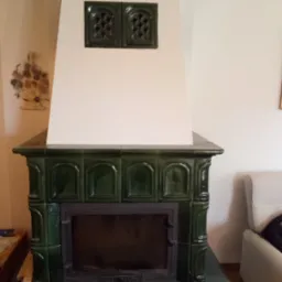 Zabudowany, zielony piec kaflowy z czarnymi drzwiczkami i ozdobnymi kratkami wentylacyjnymi w górnej części, na tle białej ściany i fragmentu salonu.