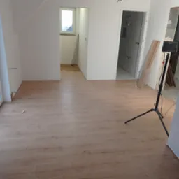 Nowo ułożona podłoga z paneli w jasnym odcieniu drewna, widoczne fragmenty łazienki z szarą glazurą, lampa budowlana na trójnogu i biały regał modułowy, okno w tle.