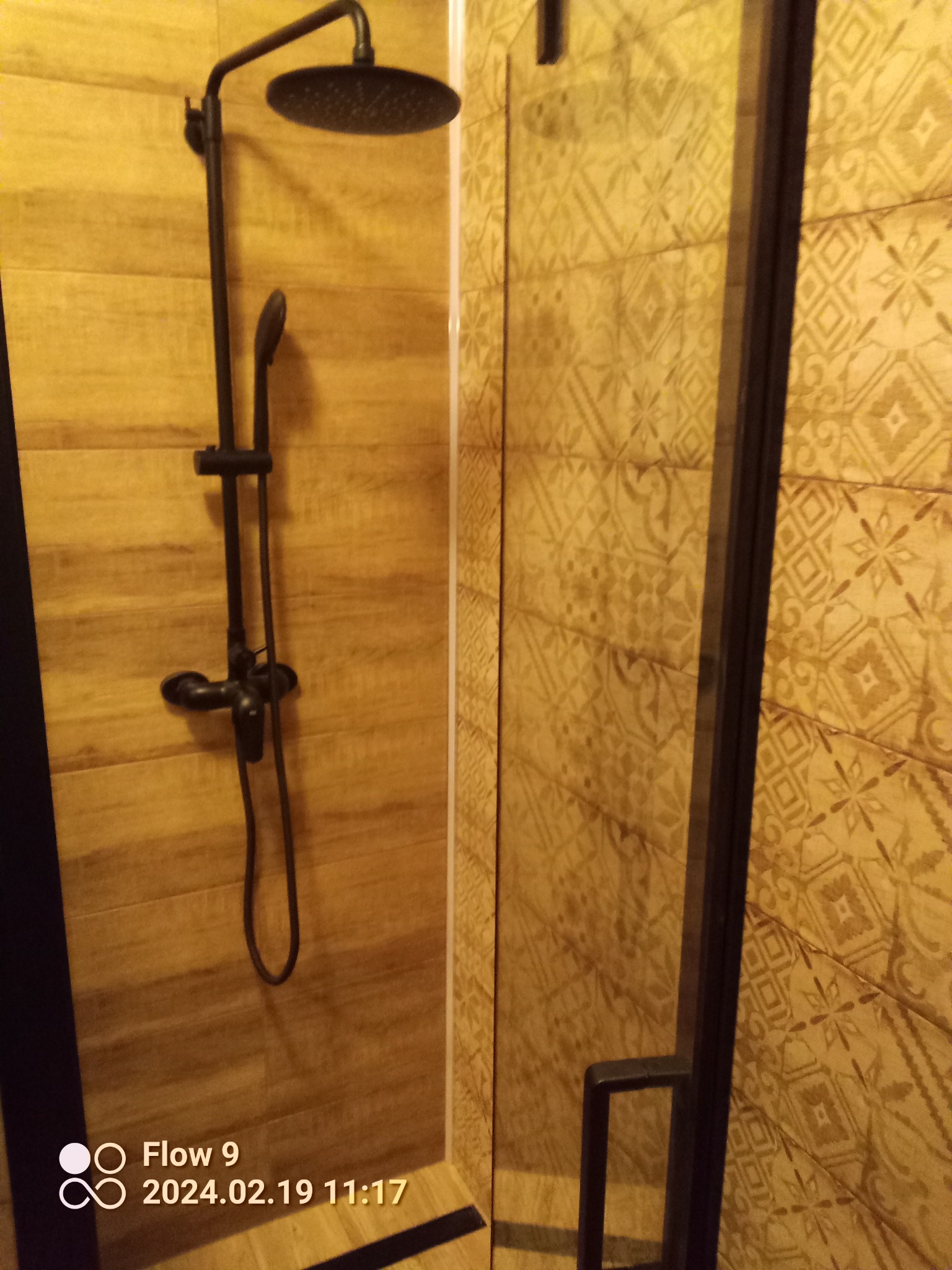 Kabina prysznicowa z czarną armaturą i dekoracyjnymi płytkami w odcieniach beżu i brązu, widoczny fragment szklanej ściany z czarną ramą.