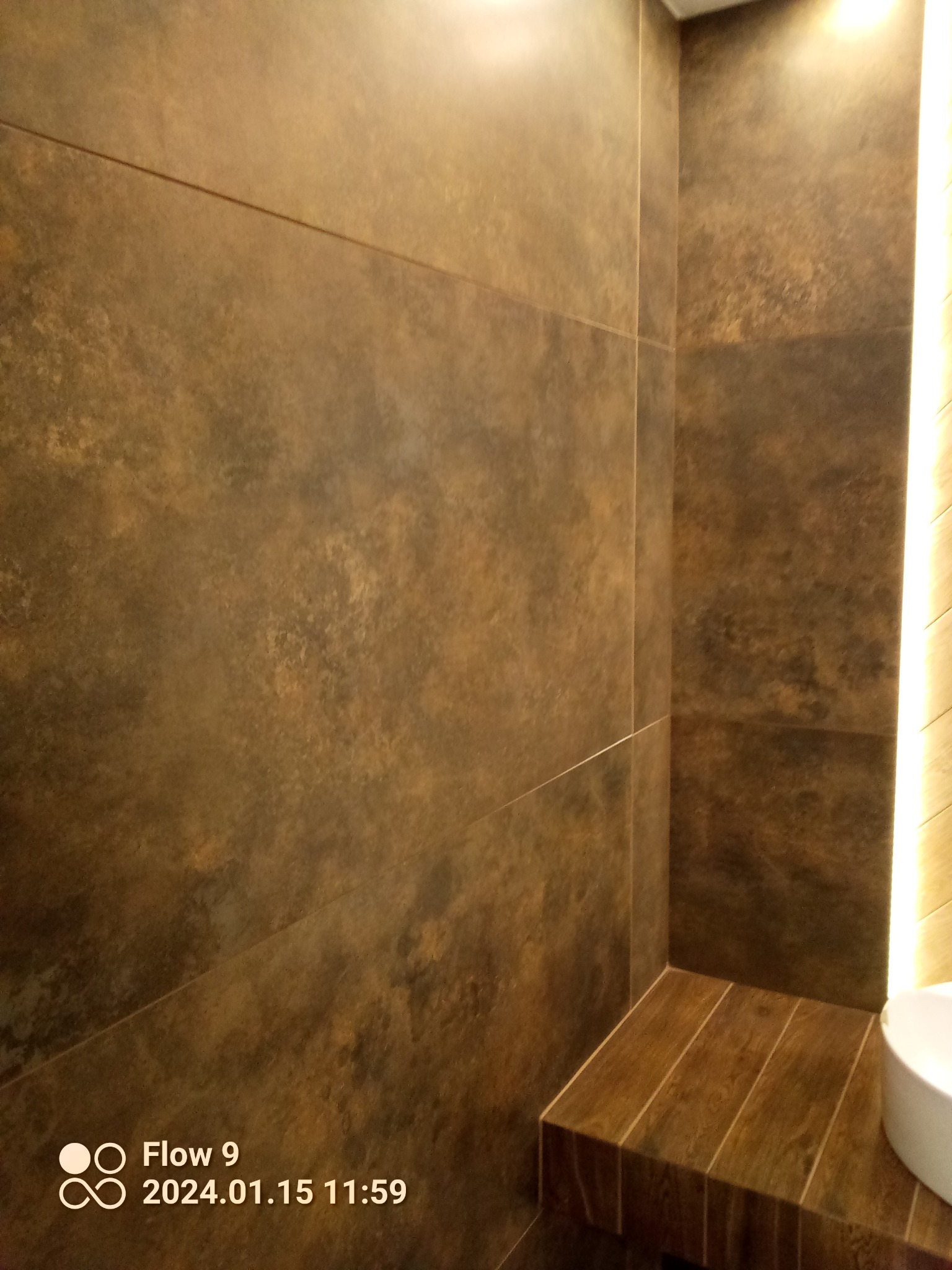 Brązowe, matowe płytki imitujące kamień na ścianach łazienki, z widoczną krawędzią zabudowy i fragmentem białej umywalki, oświetlone pionowym pasem LED.