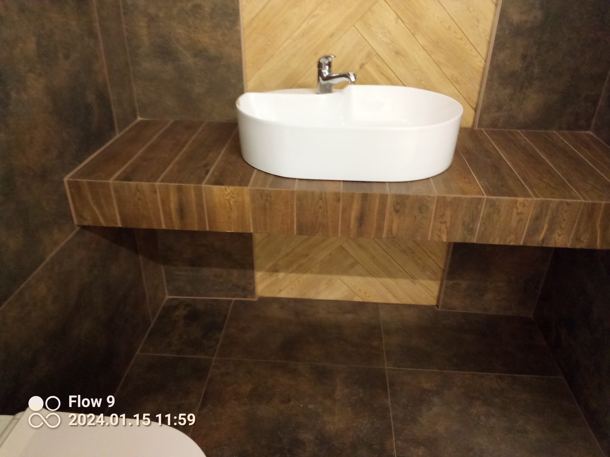 Umywalka nablatowa z chromowaną baterią na blacie z płytek imitujących drewno, brązowe płytki na ścianach i podłodze, wzór jodełki na ścianie za umywalką.