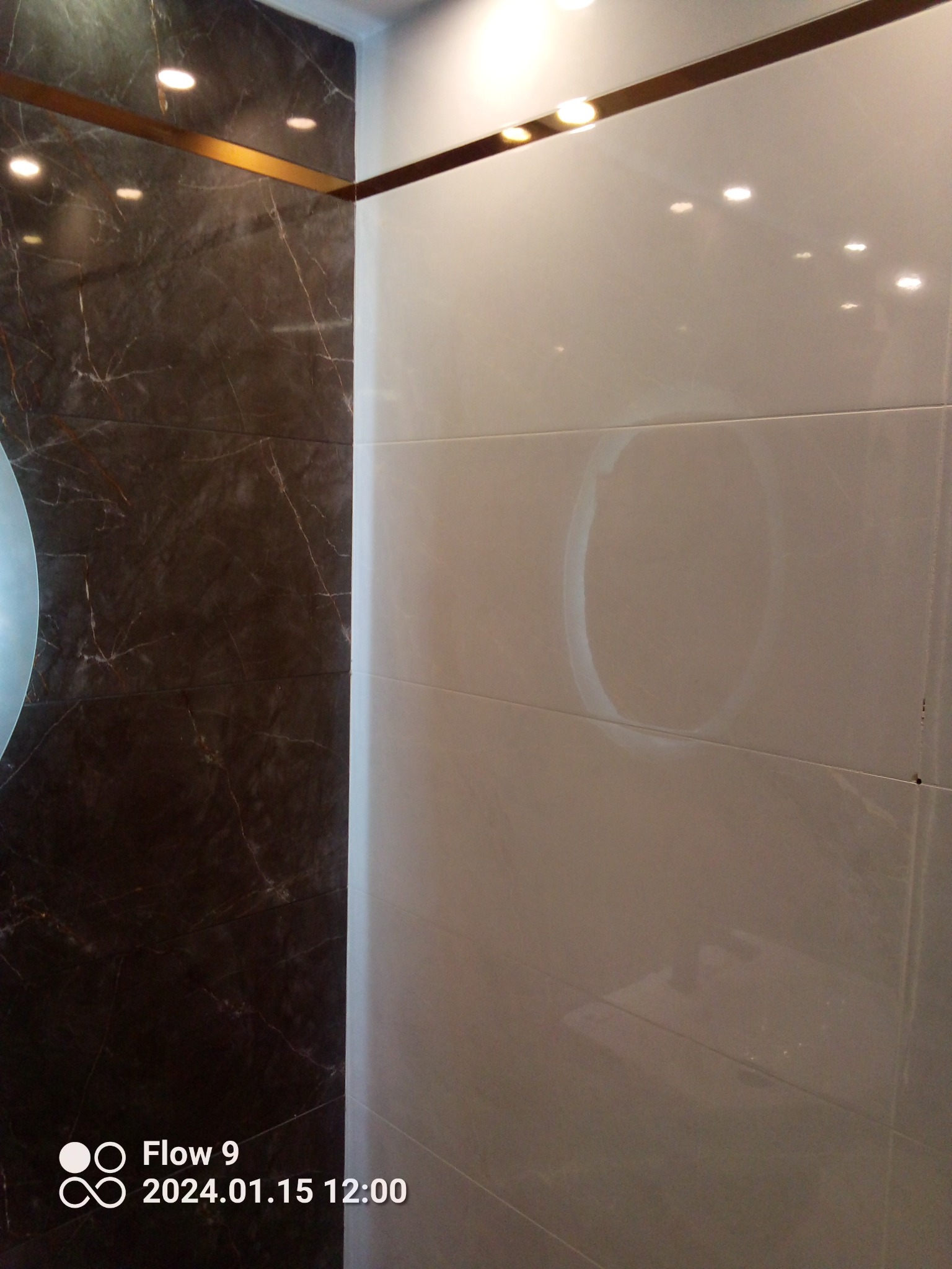 Wykończona ściana łazienki z ciemnymi, marmurkowymi płytkami po lewej i jasnymi, błyszczącymi płytkami po prawej stronie, oddzielonymi złotą listwą dekoracyjną.