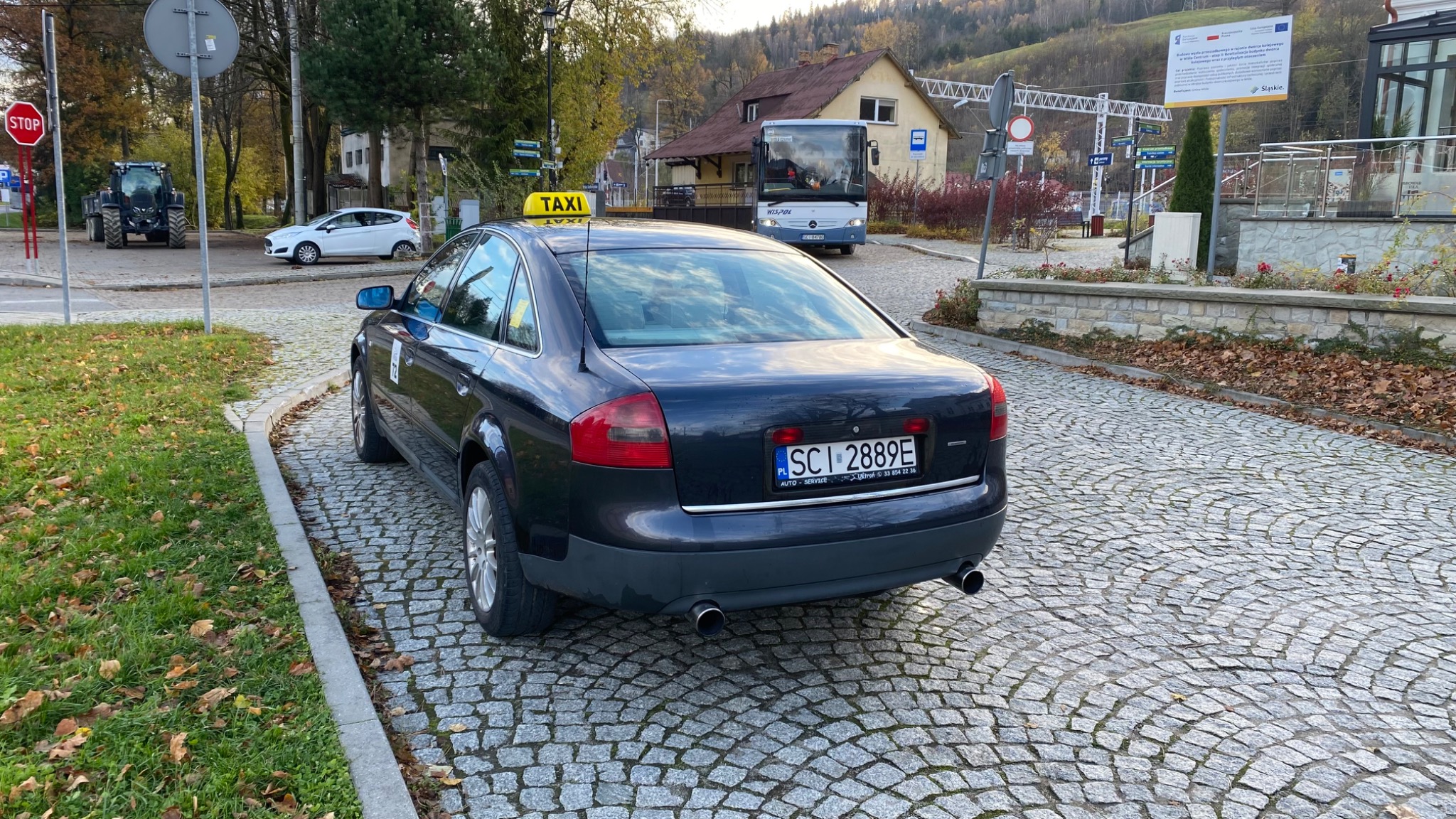 Czarny samochód osobowy z żółtym napisem TAXI na dachu, zaparkowany na brukowanej ulicy w Wiśle, w tle budynek stacji kolejowej i autobus.