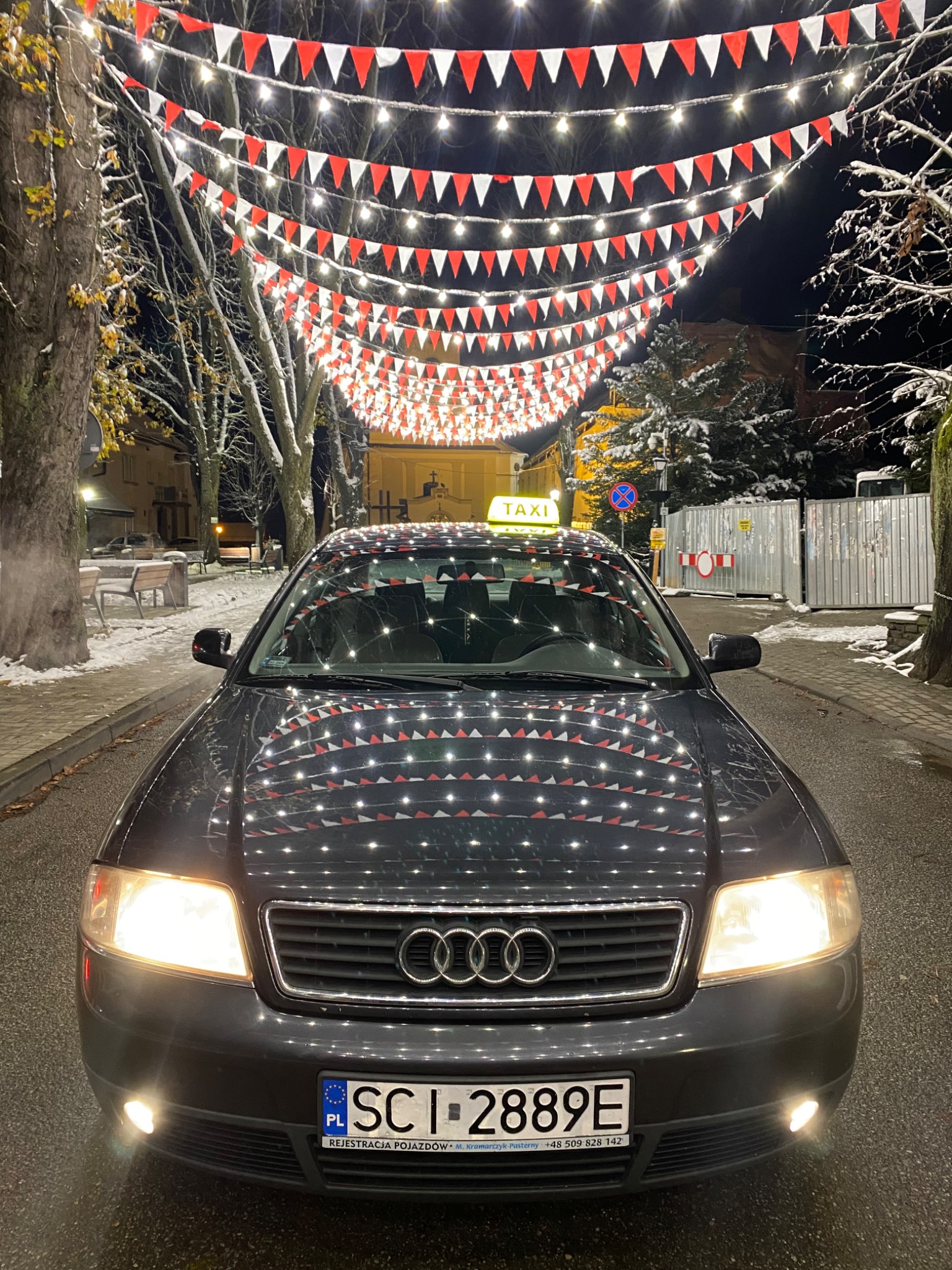 Ciemnoszara taksówka Audi A6 z zapalonymi światłami, tablicą rejestracyjną SC I 2889E i lampą TAXI na dachu, sfotografowana nocą na tle ulicy udekorowanej czerwono-białymi flagami i lampkami.