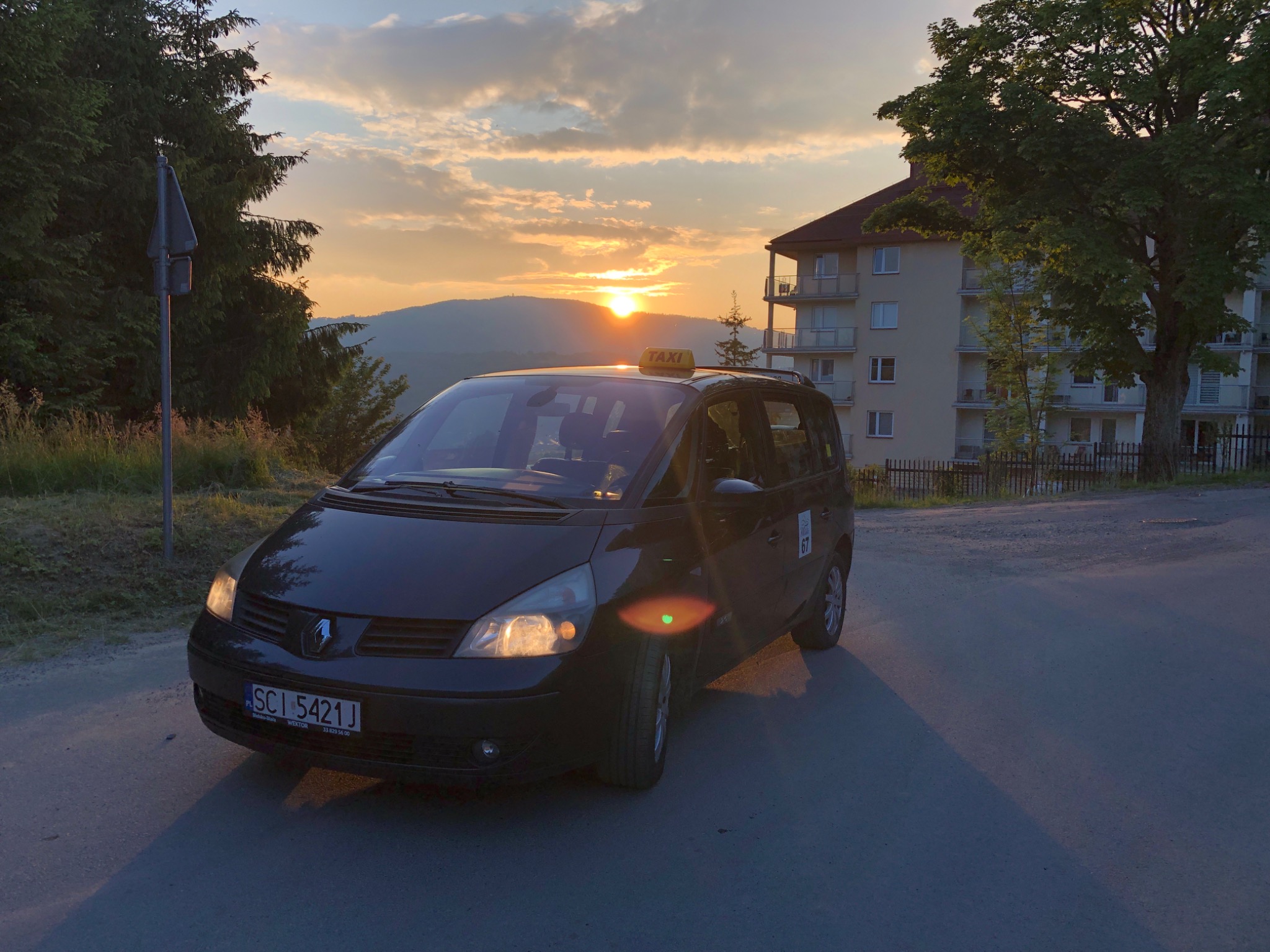 Czarny van taxi Renault Espace na tle zachodzącego słońca i górskiego krajobrazu, z widocznym znakiem taxi na dachu i numerem rejestracyjnym SCI 5421J.