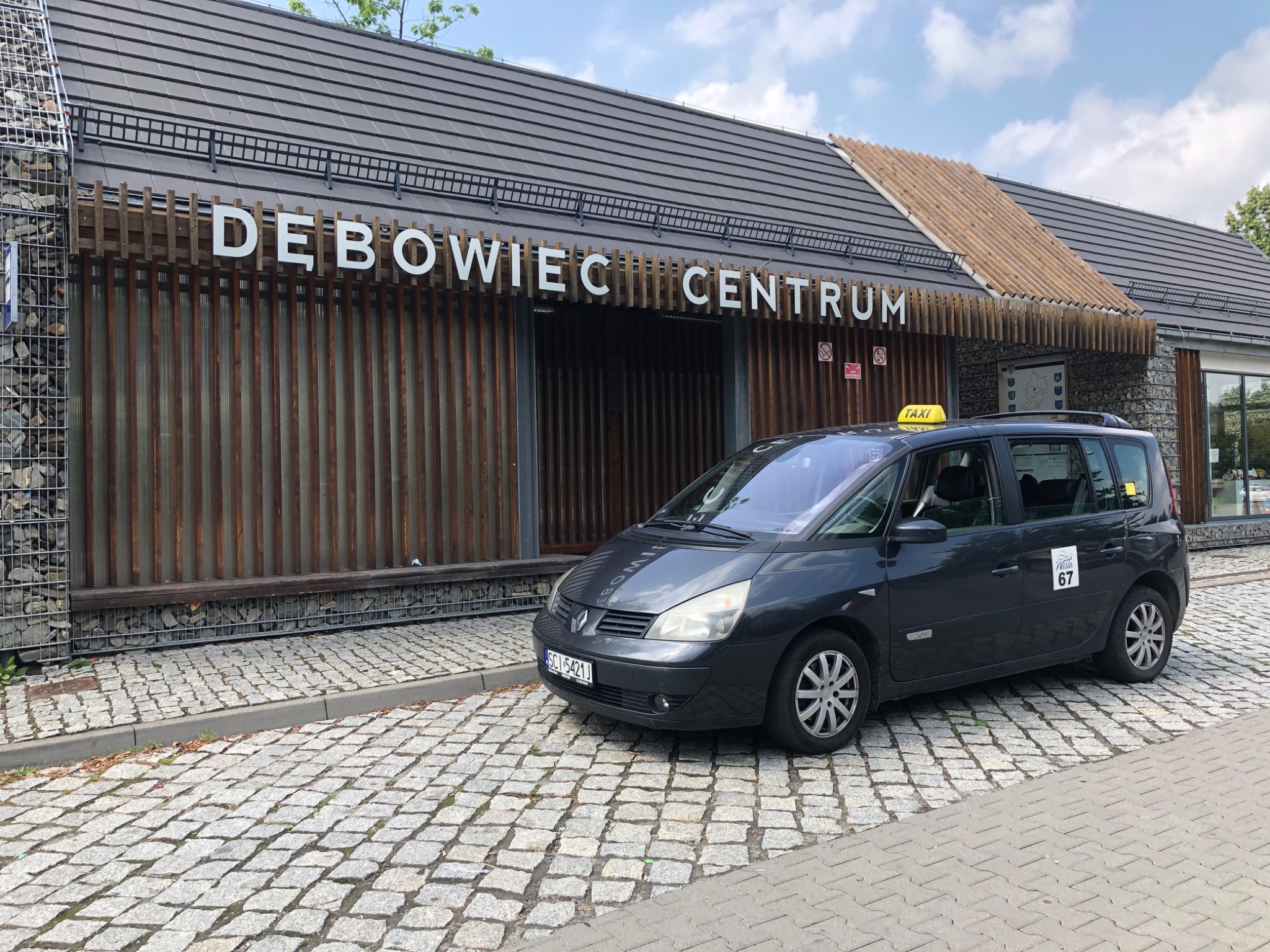 Ciemnoszary minivan taxi parkuje przed budynkiem Dębowiec Centrum z drewnianą fasadą i białym napisem, na żółtym dachu taksówki widoczny napis TAXI.