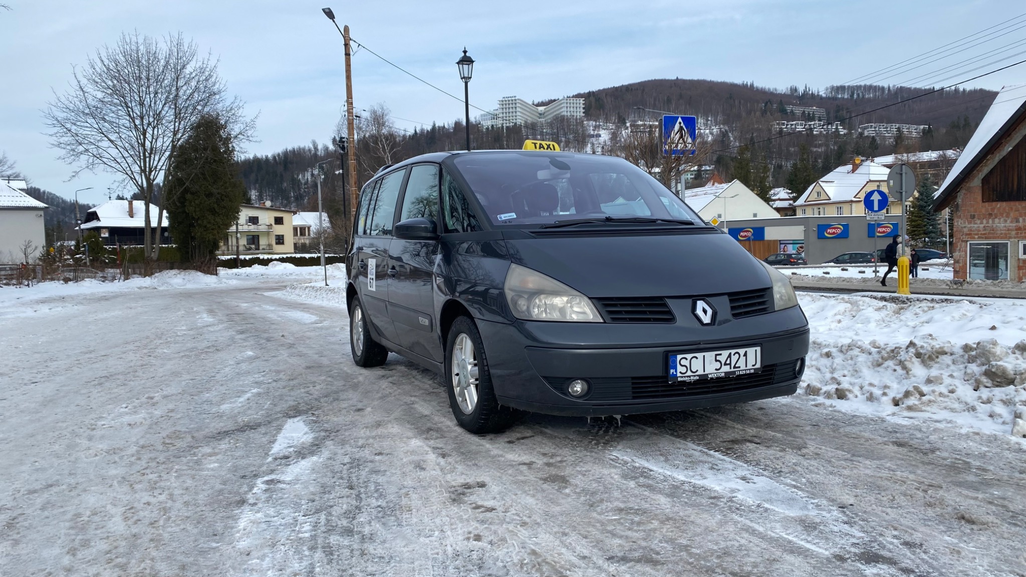 Ciemnoszary Renault Espace z żółtym szyldem TAXI na dachu, zaparkowany na oblodzonej drodze w zimowej scenerii górskiego miasteczka.