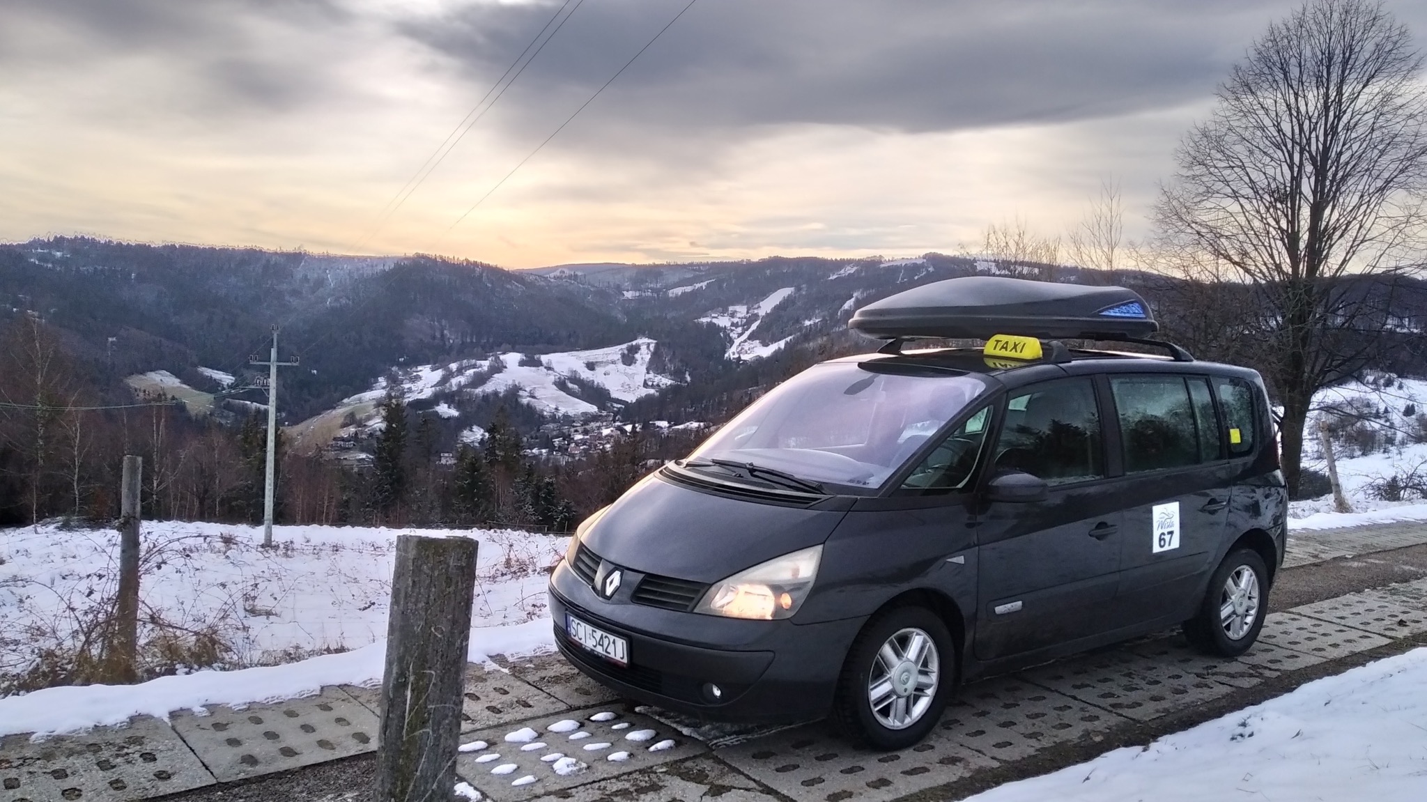 Czarny van Renault Espace z żółtym szyldem TAXI na dachu, wyposażony w bagażnik dachowy, zaparkowany na tle zimowego krajobrazu górskiego z widocznymi ośnieżonymi wzgórzami i zachodzącym słońcem.