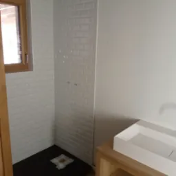 Nowoczesna łazienka w trakcie remontu: białe kafelki typu metro na ścianach kabiny prysznicowej, czarny brodzik zabezpieczony folią, drewniana rama okienna i szafka pod umywalkę z blatem.