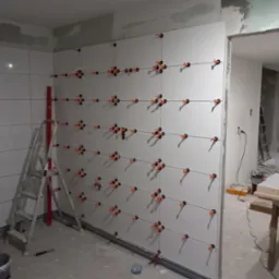 Świeżo ułożone, białe płytki na ścianie w trakcie fugowania, z użyciem systemu poziomowania płytek (klipsy i kliny), widoczna drabina i poziomica.