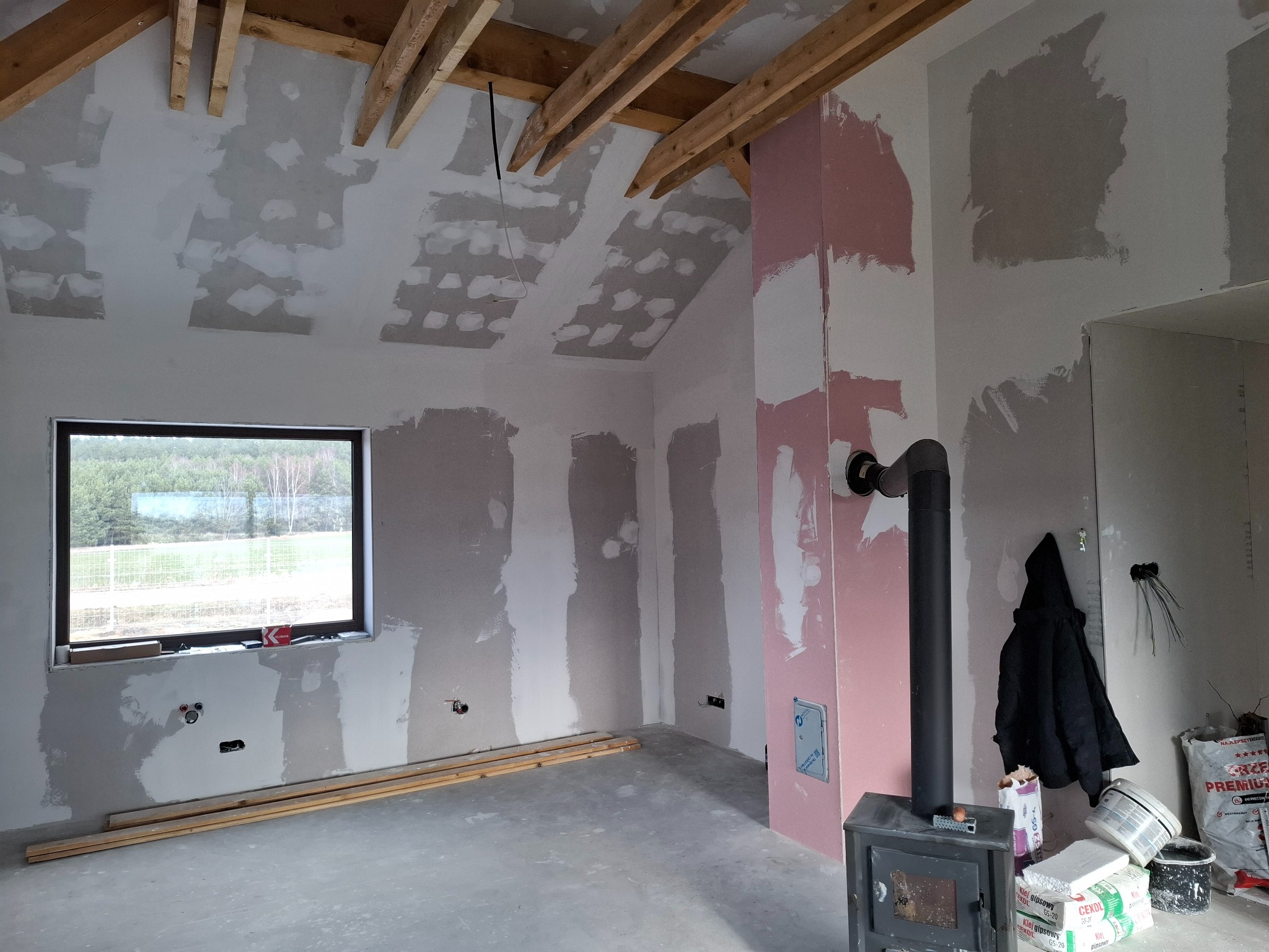Wnętrze w trakcie remontu: surowy stan ścian z widocznymi łatami gipsowymi, odsłonięta konstrukcja drewniana dachu, piec wolnostojący z czarną rurą, okno z widokiem na zieleń, worki z zaprawą...