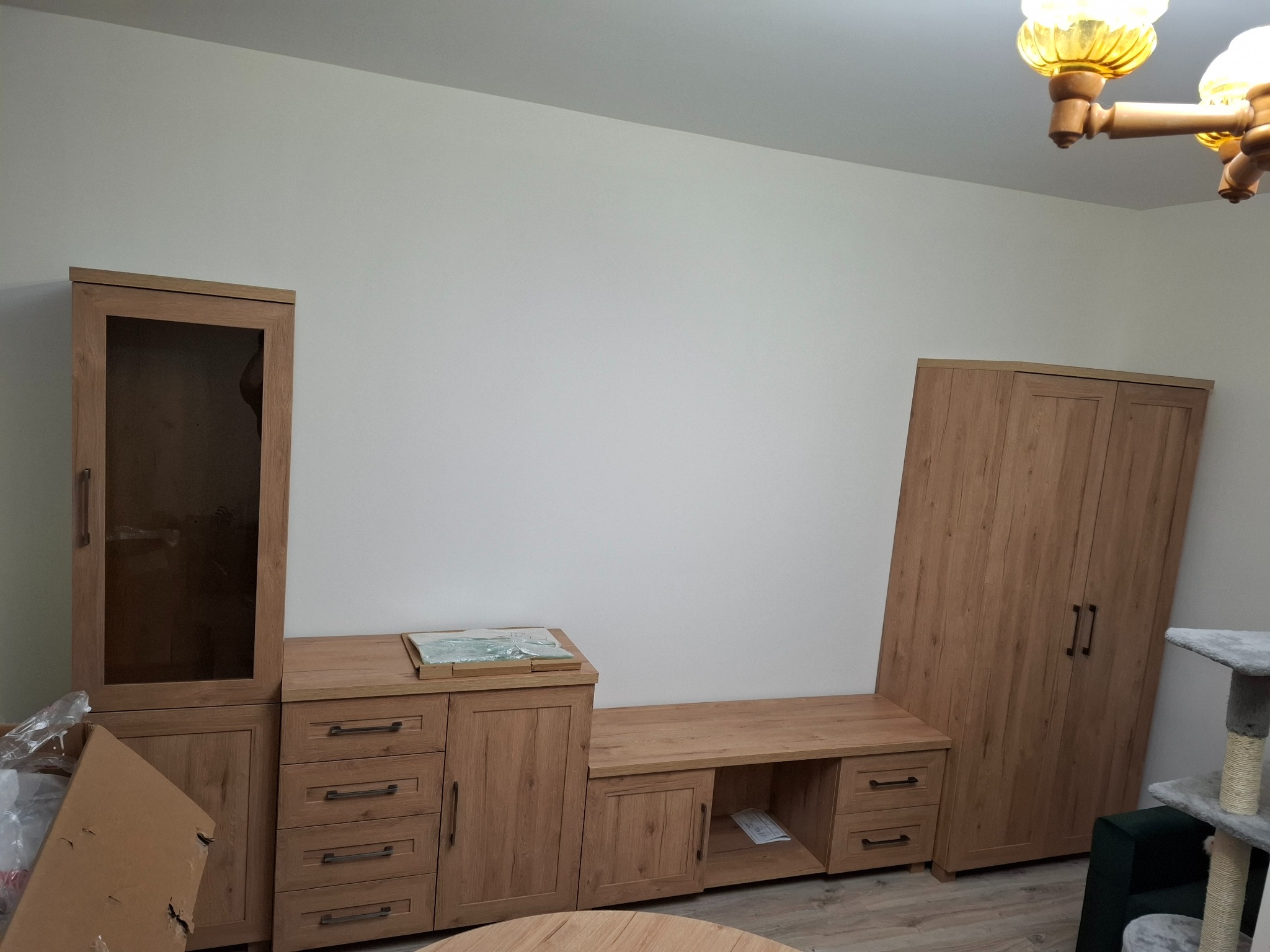 Częściowo zmontowany zestaw mebli w odcieniach dębu, z widocznymi elementami w kartonowym opakowaniu, ustawiony w pokoju z kremowymi ścianami i drewnianą podłogą.