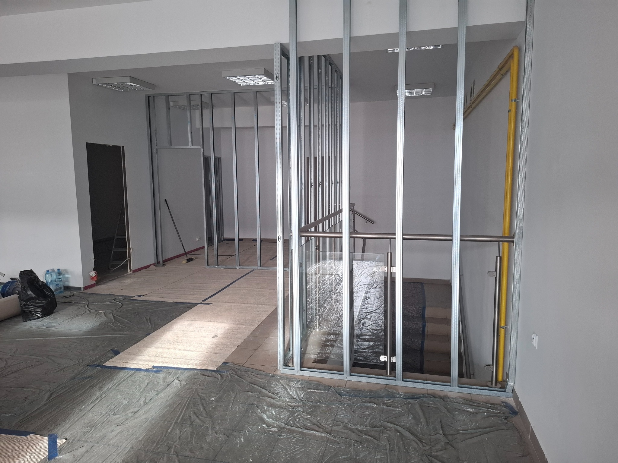Wnętrze w trakcie remontu: widoczne metalowe profile konstrukcyjne ścian działowych, zabezpieczona folią podłoga, fragment schodów z balustradą oraz żółta rura instalacyjna przy ścianie.