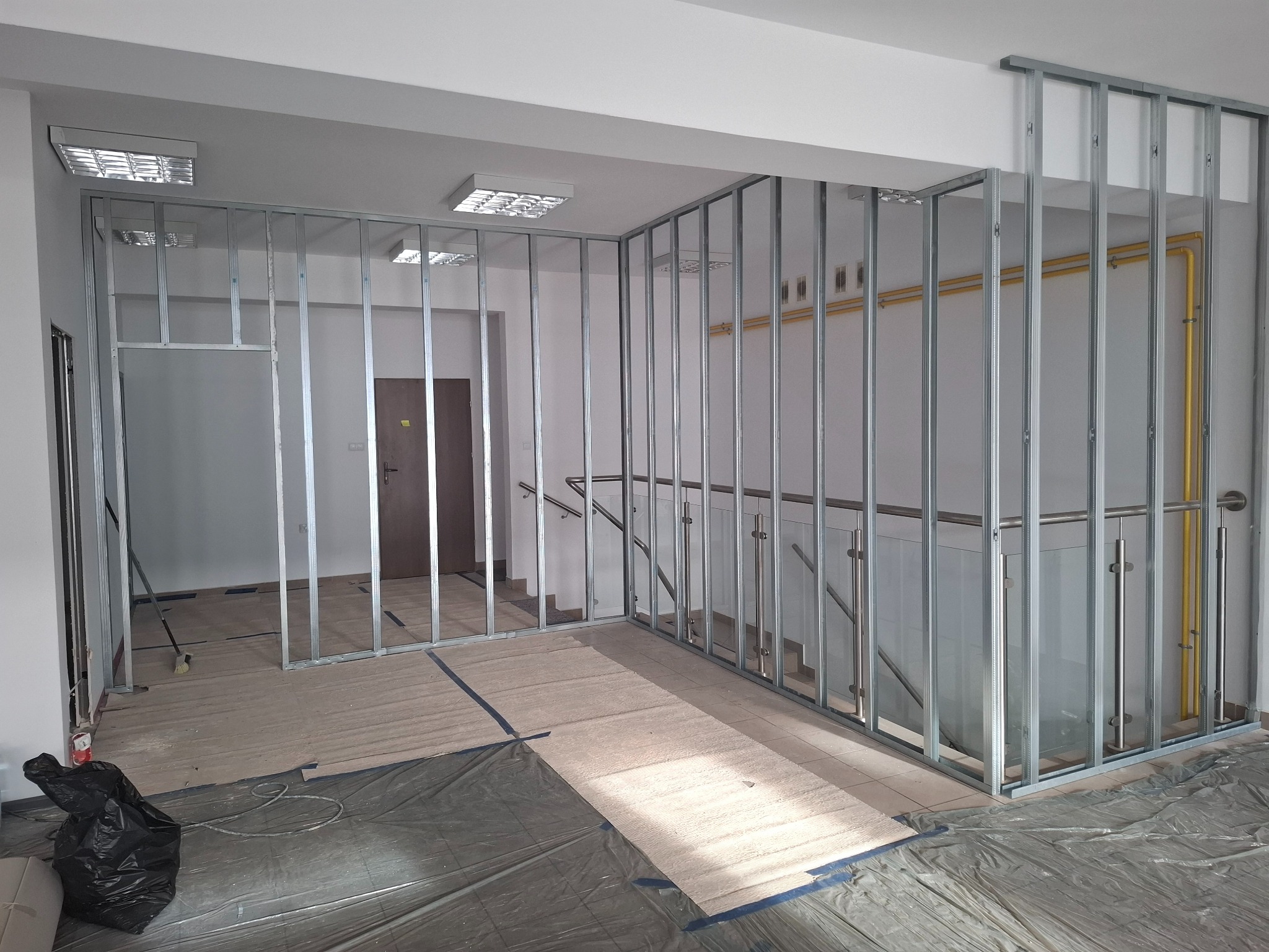 Metalowa konstrukcja szkieletowa ścianki działowej w trakcie montażu, widoczne rury instalacyjne w tle, zabezpieczona podłoga folią i kartonem.