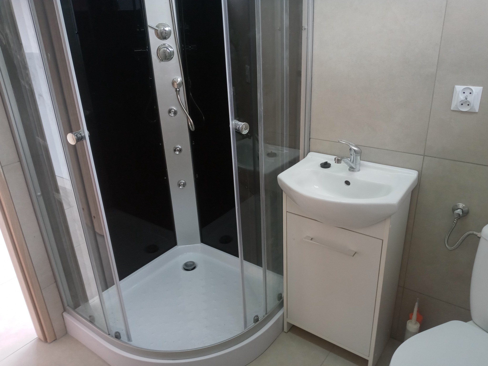 Nowoczesna kabina prysznicowa z czarnym panelem i chromowanymi elementami, umywalka z szafką w minimalistycznej łazience z jasnymi płytkami.