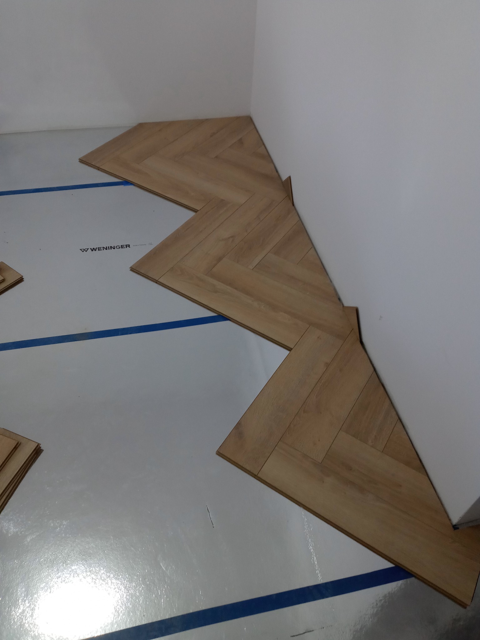 Układanie paneli podłogowych w jodełkę, widoczne przycięte panele przy ścianie, folia izolacyjna Wenninger z niebieskimi liniami pomocniczymi.