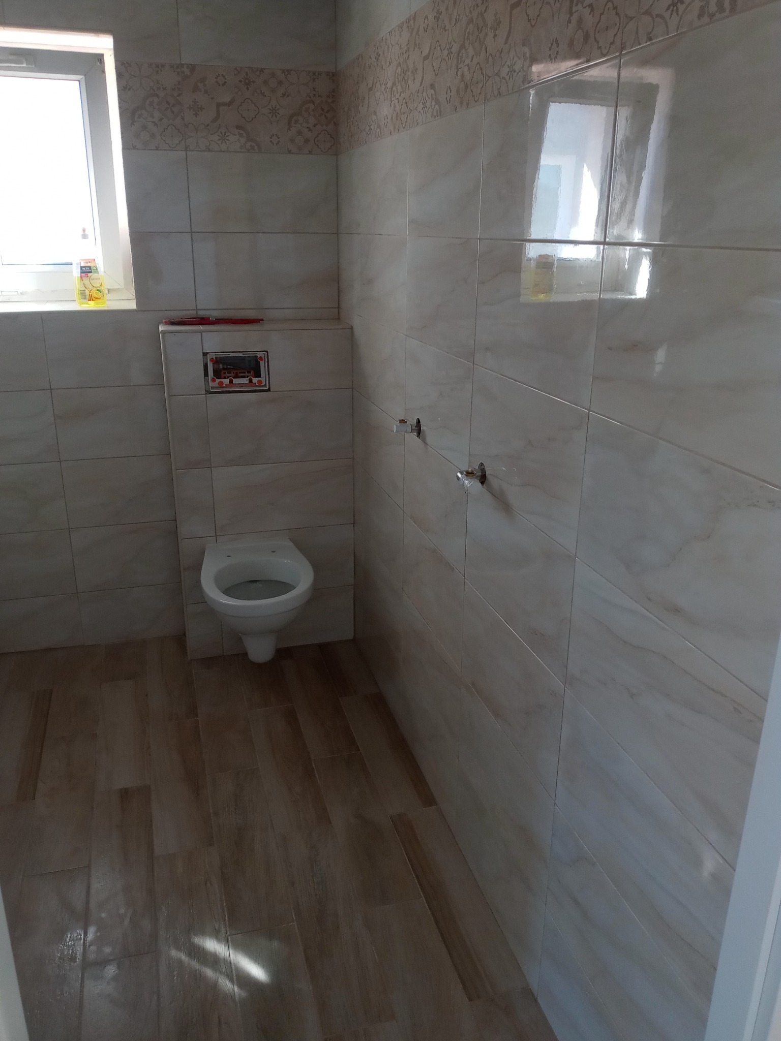 Wykończona łazienka z beżowymi płytkami imitującymi marmur na ścianach i podłodze, wisząca toaleta z zabudowaną spłuczką, okno z widocznym płynem do mycia.