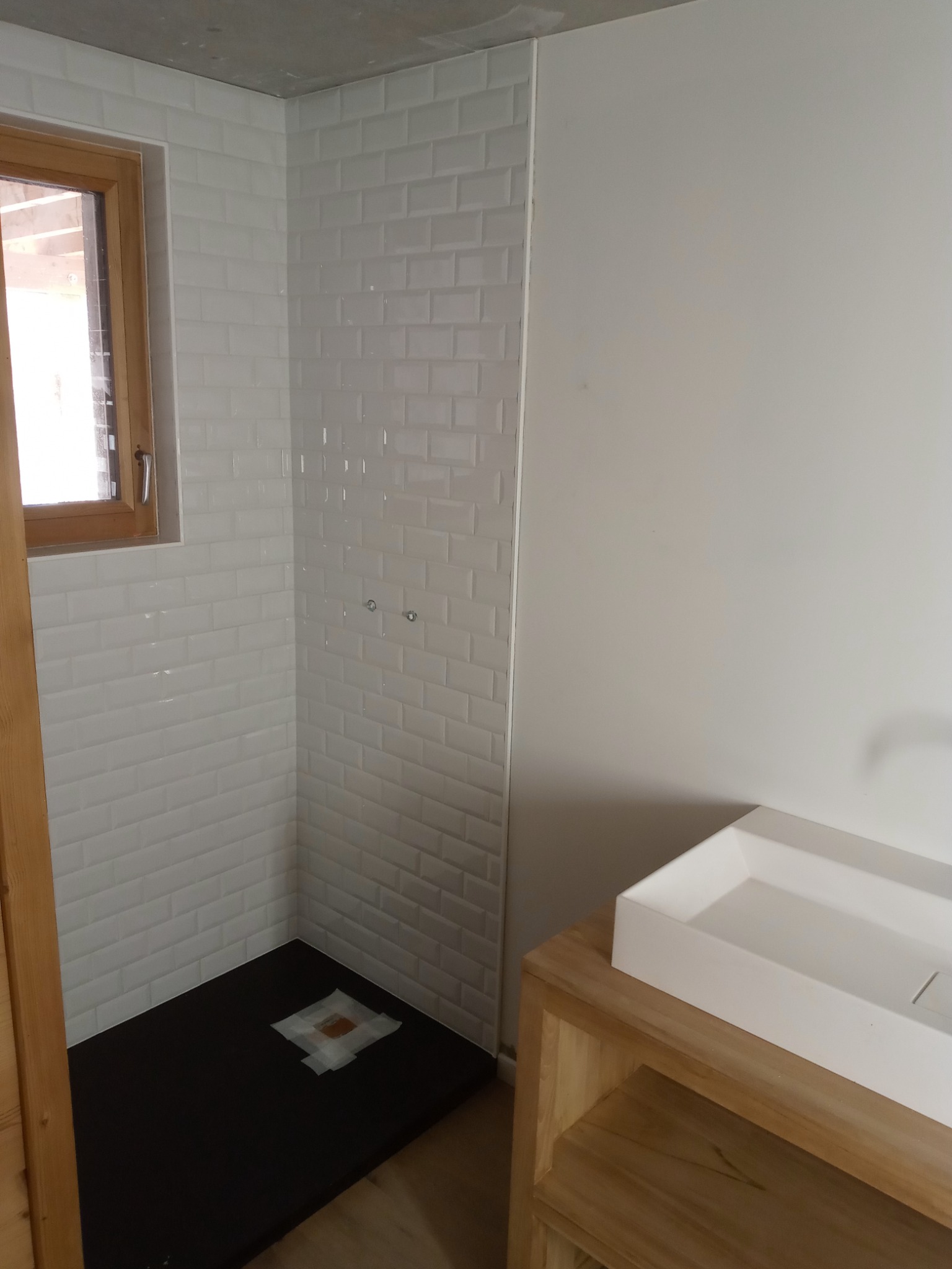 Nowoczesna łazienka w trakcie remontu: białe kafelki typu metro na ścianach kabiny prysznicowej, czarny brodzik zabezpieczony folią, drewniana rama okienna i szafka pod umywalkę z blatem.