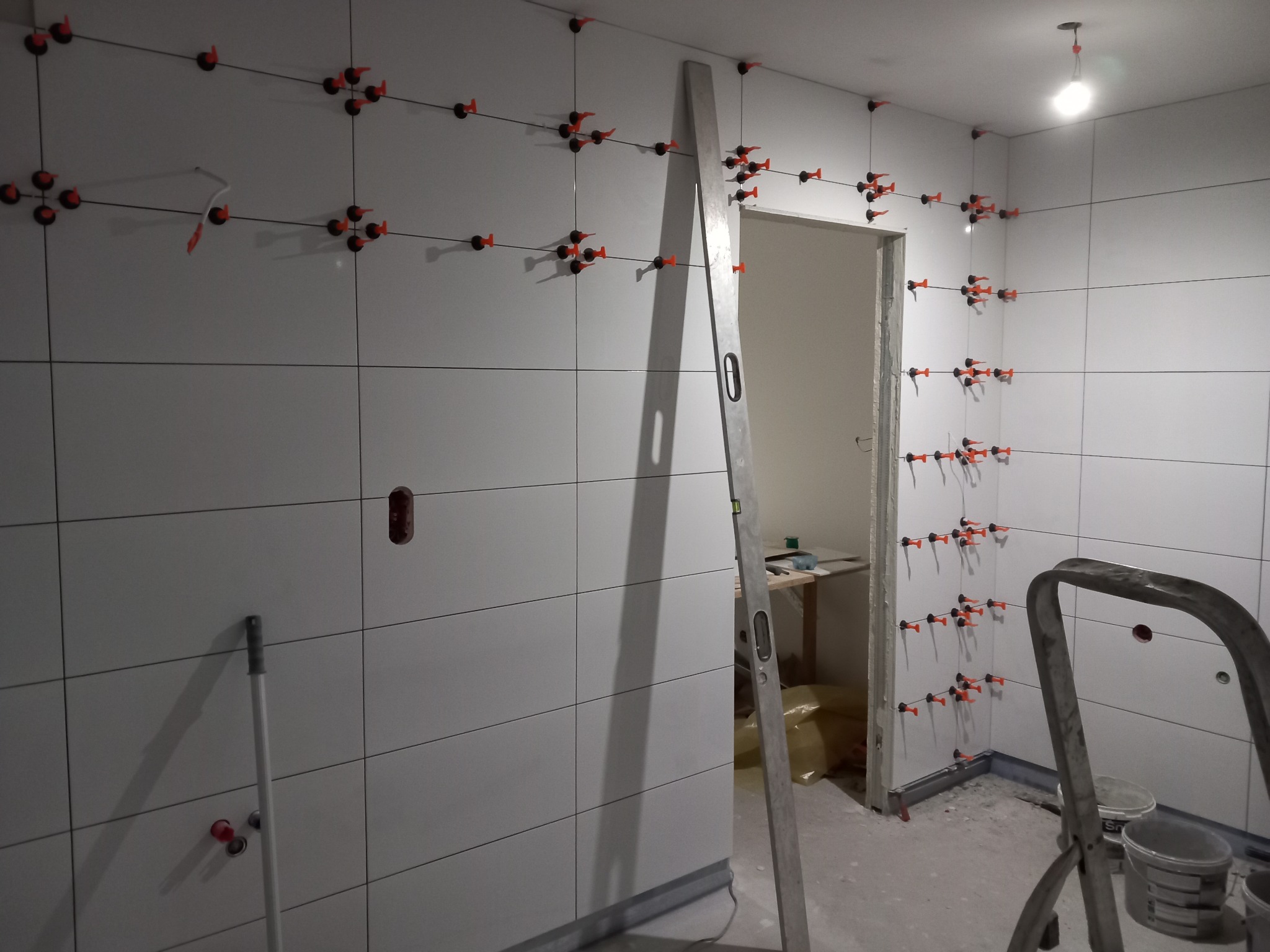 Świeżo ułożone, białe płytki ceramiczne na ścianach łazienki, z użyciem poziomujących klipsów i krzyżyków, widoczna poziomica oparta o ścianę i drabina