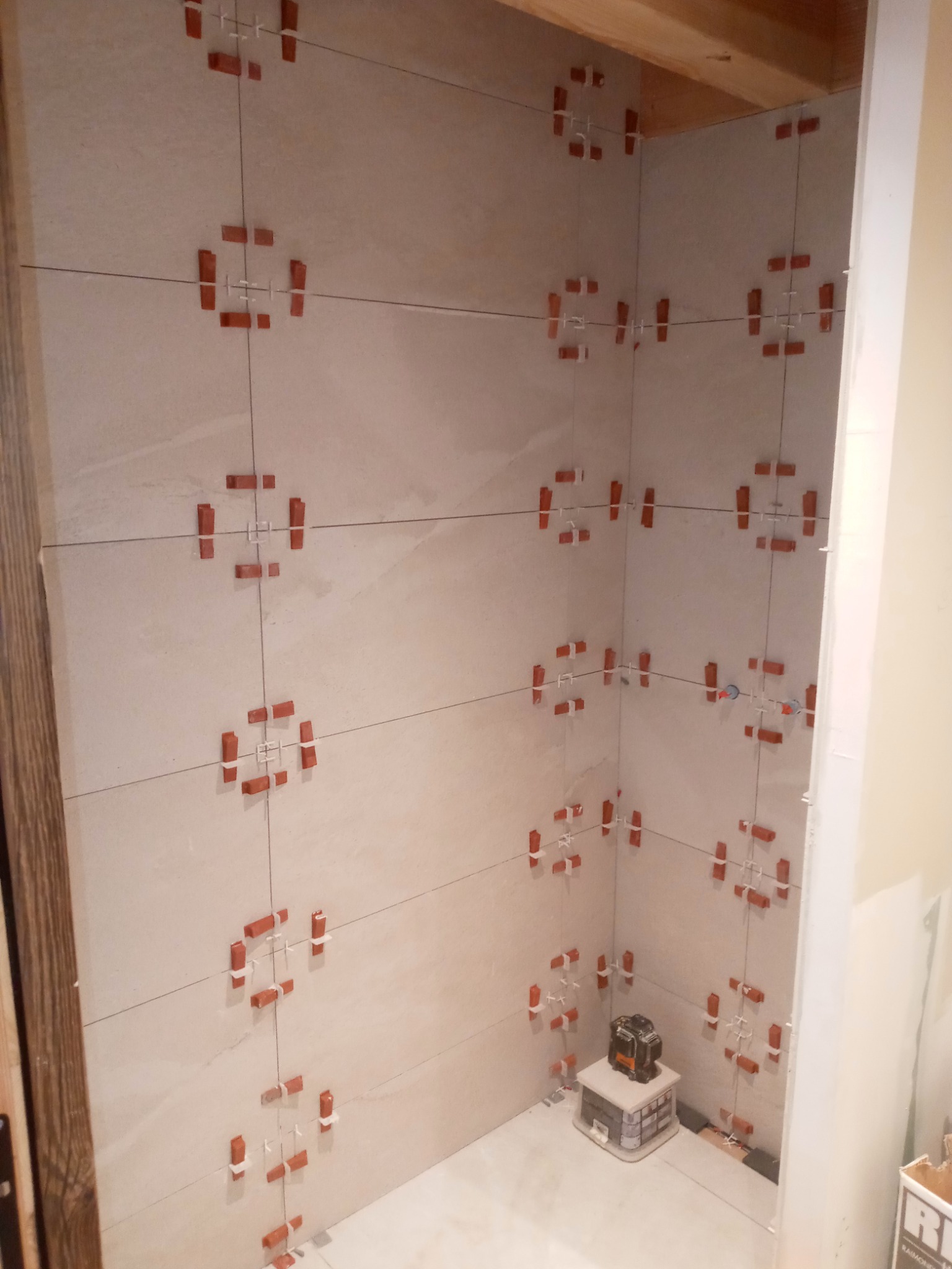 Ściany łazienki w trakcie układania jasnych płytek, widoczne krzyżyki dystansowe i laserowy niwelator na podłodze.
