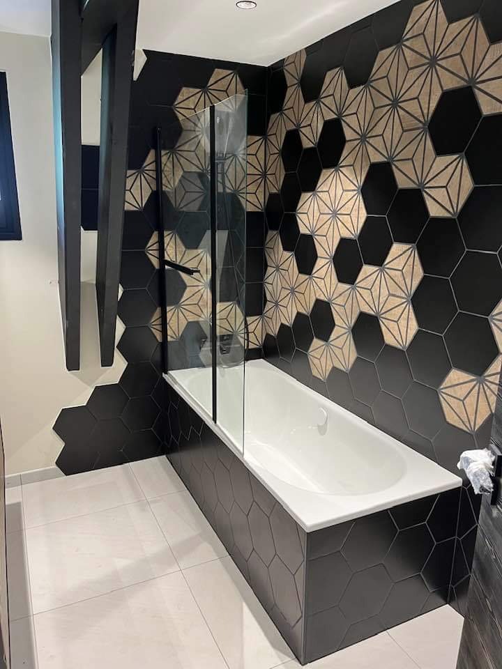Nowoczesna łazienka z wanną obudowaną czarnymi, heksagonalnymi płytkami, szklany parawan nawannowy, ściana z dekoracyjnymi płytkami w geometryczne wzory, biała podłoga.