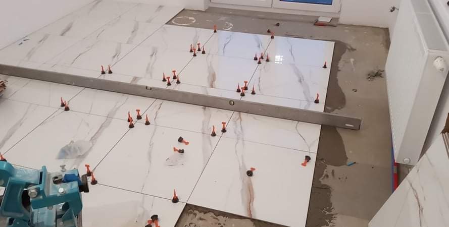 Układanie białych płytek imitujących marmur na podłodze, z użyciem poziomicy i czerwonych klinów do poziomowania.