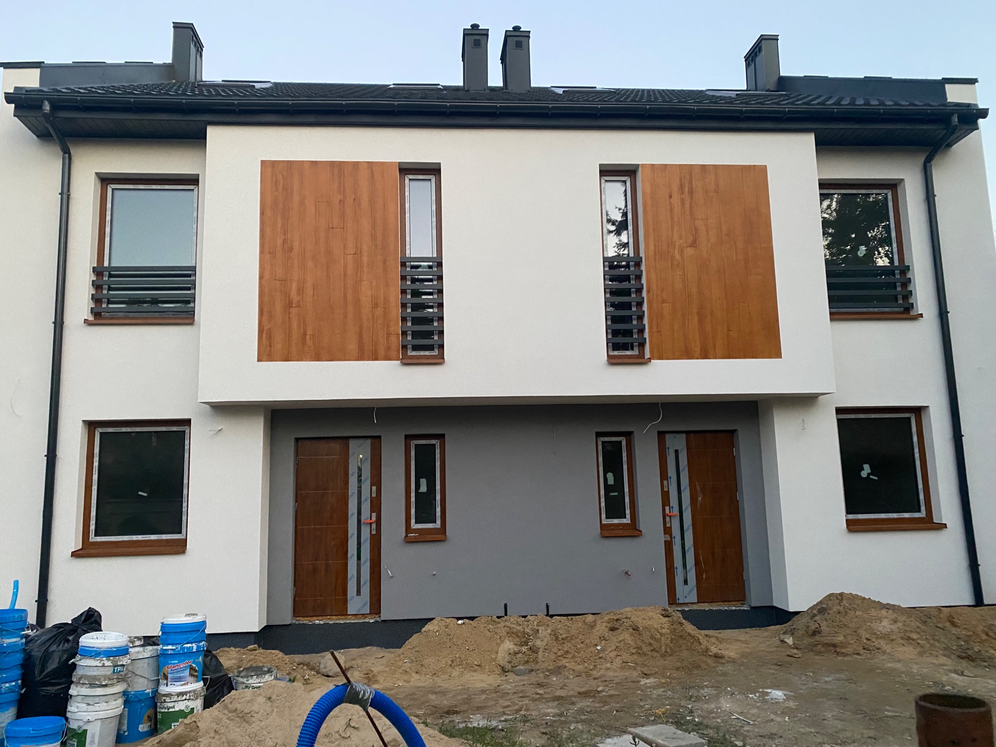 Bliźniak z białą elewacją, drewnianymi panelami okiennymi i szarą strefą wejściową. Widoczne wiaderka z farbą i piasek sugerują prace wykończeniowe.