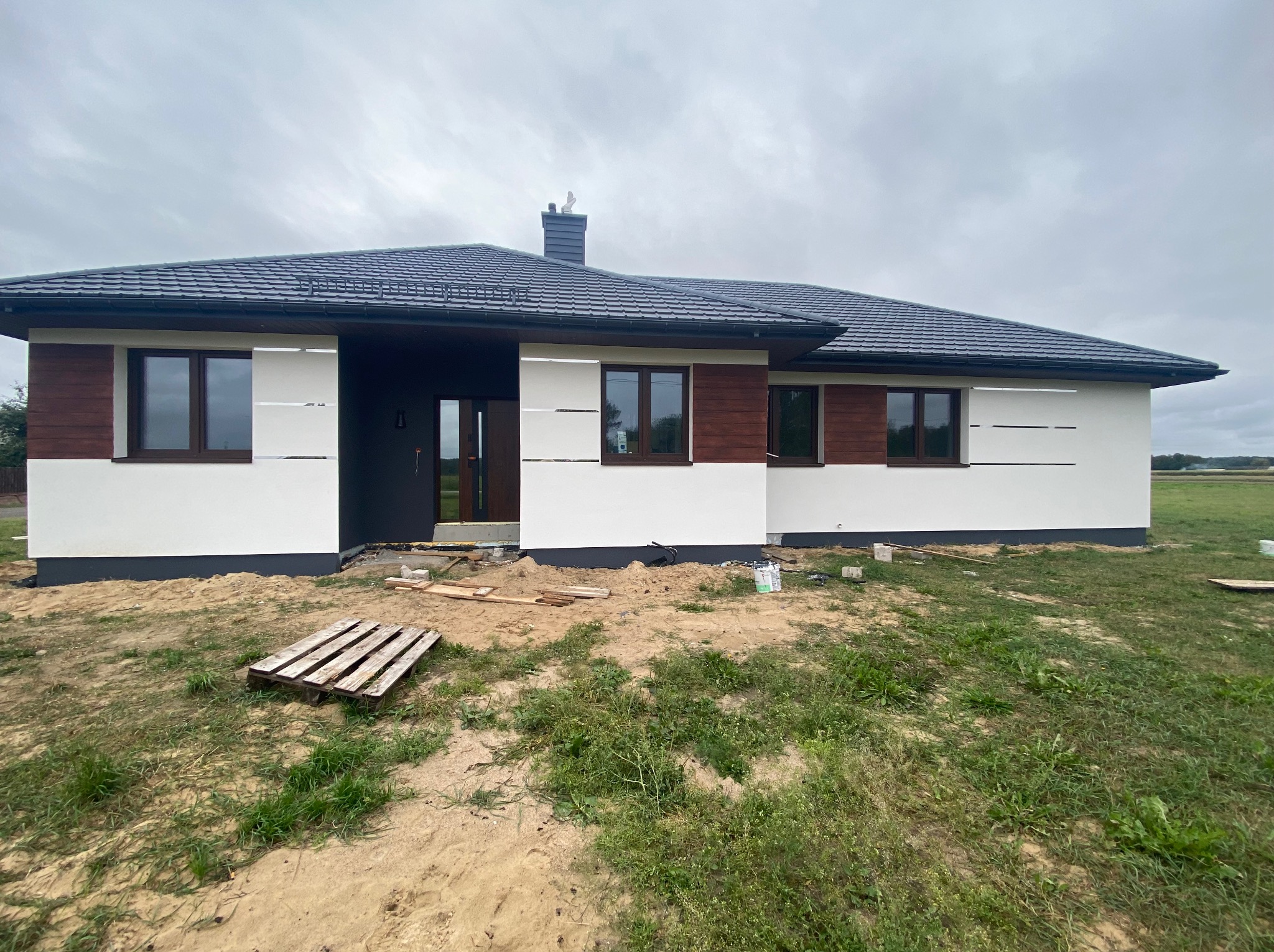 Nowoczesny dom parterowy z ciemną dachówką i elementami drewnianymi na elewacji. Widoczne niedokończone prace ziemne wokół budynku i paleta budowlana.