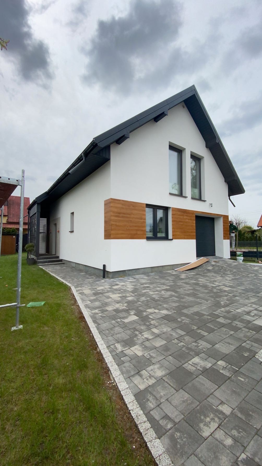 Nowoczesny dom jednorodzinny z białą elewacją, drewnianymi akcentami i szarą kostką brukową na podjeździe. Widoczne elementy wykończenia i świeżo założony trawnik.