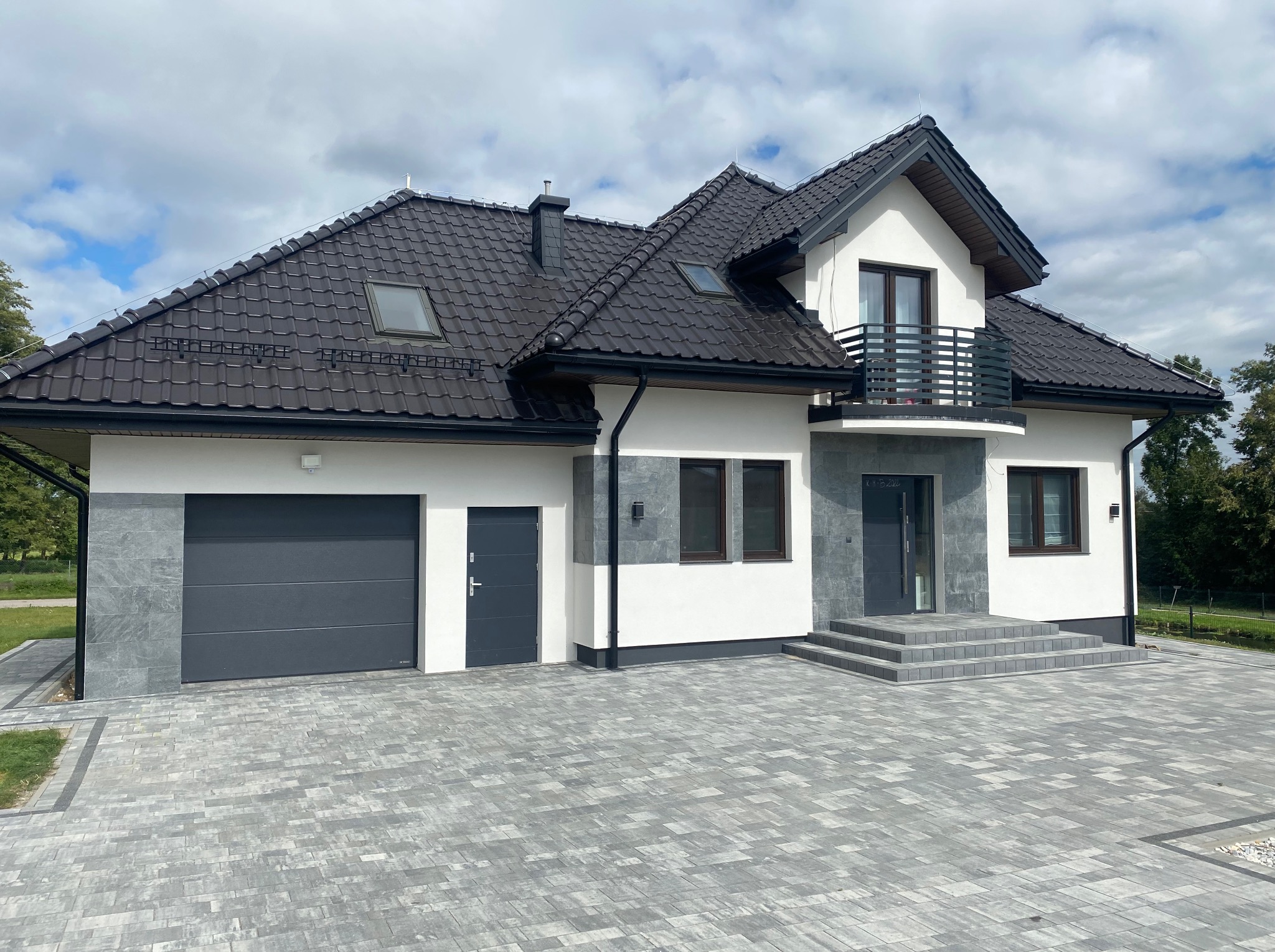 Nowoczesny dom jednorodzinny z balkonem, wykończony w bieli i szarości, z grafitową stolarką i dachem. Uwagę zwraca elewacja z elementami kamiennymi i kostka brukowa.