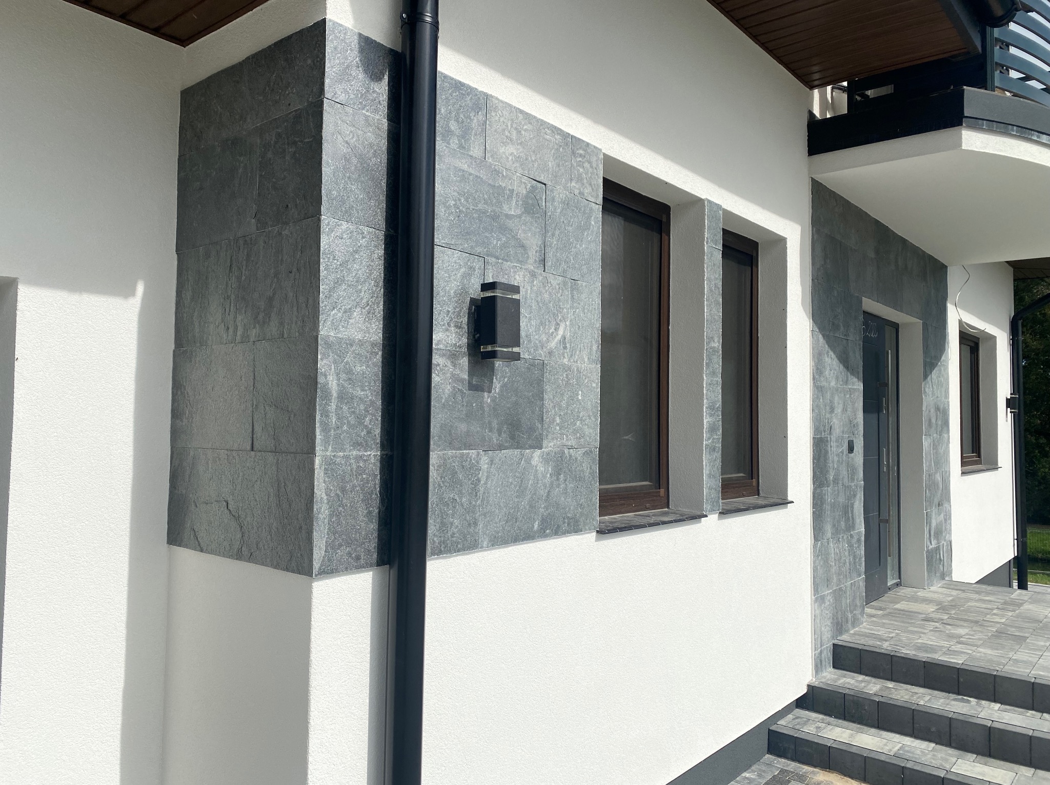 Wykończona elewacja domu z szarej płytki imitującej kamień, biały tynk, brązowe okna i czarna rynna. Nowoczesny design z oświetleniem zewnętrznym.