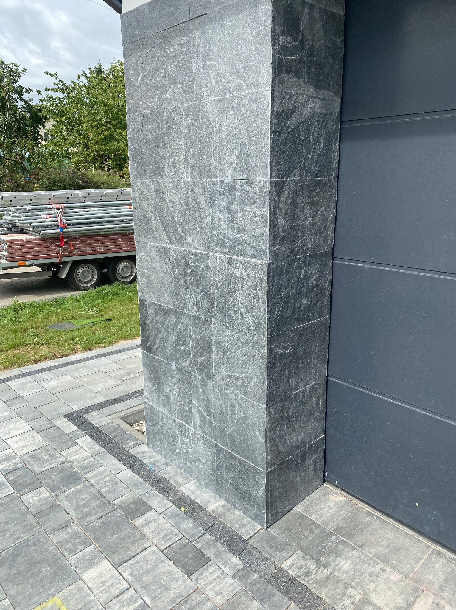 Wykończony narożnik elewacji z szarej, kamiennej okładziny, przylegający do grafitowej ściany. W tle przyczepa ze złożonym rusztowaniem i kostka brukowa.