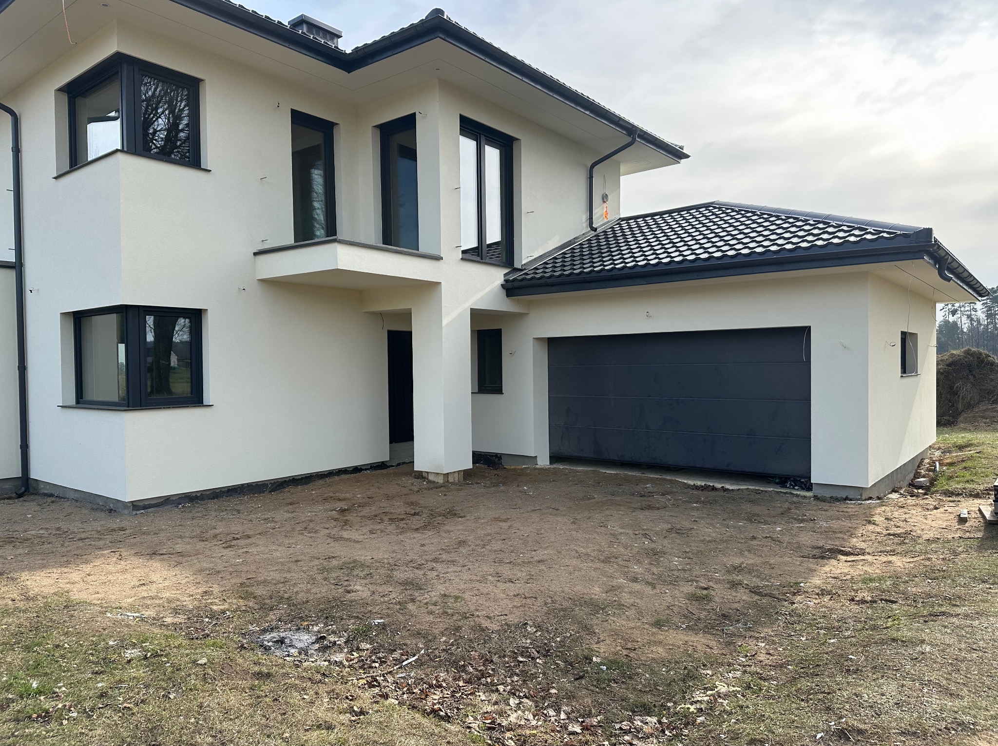 Nowoczesny dom z garażem i jasną elewacją. Minimalistyczny design, czarne ramy okienne i dachówka. Teren wokół domu przygotowany pod zagospodarowanie.
