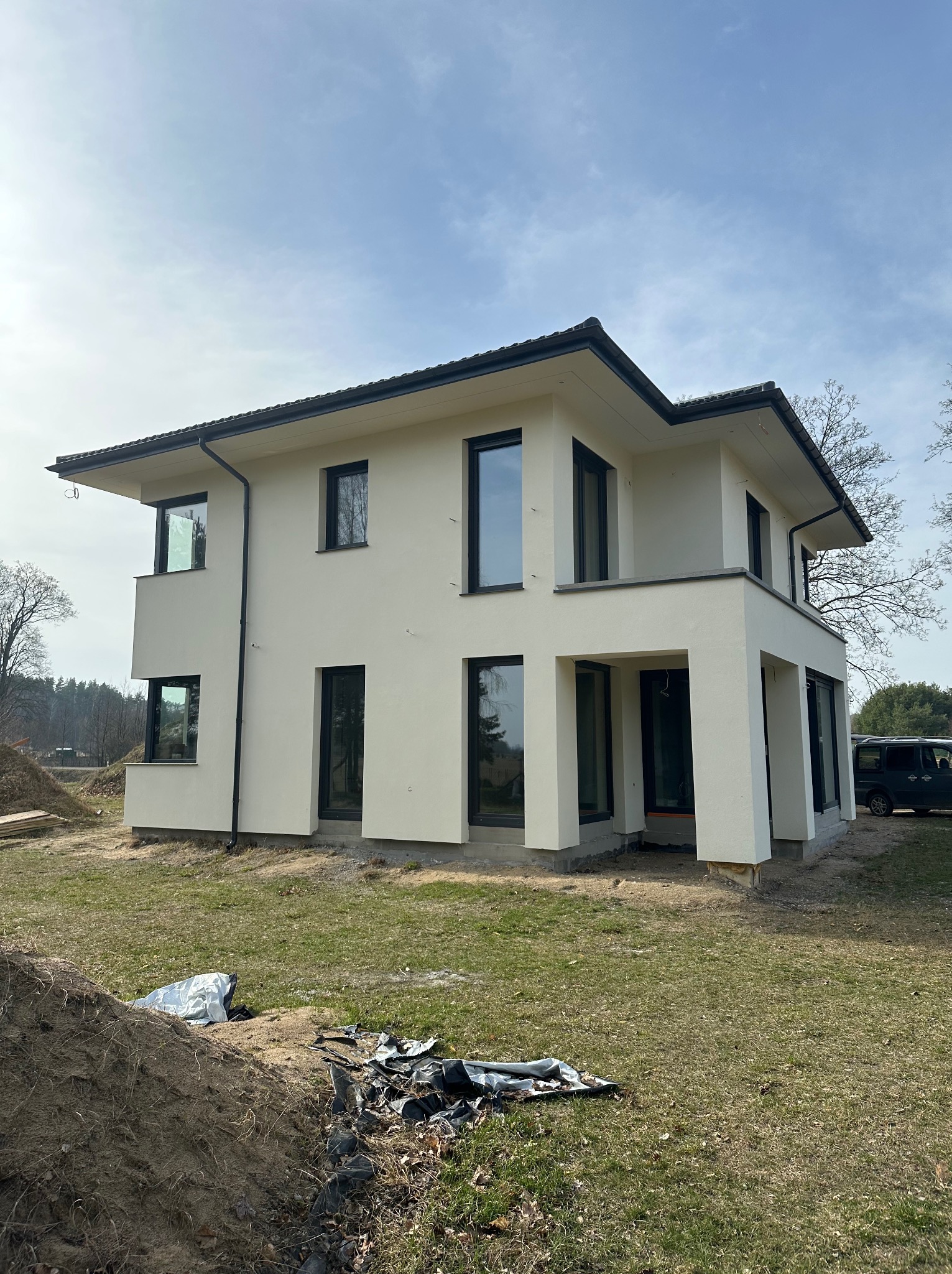 Dwukondygnacyjny dom z jasną elewacją i czarnymi ramami okiennymi, widoczny z perspektywy trawnika z elementami budowy. Minimalistyczny styl, nowoczesna architektura.