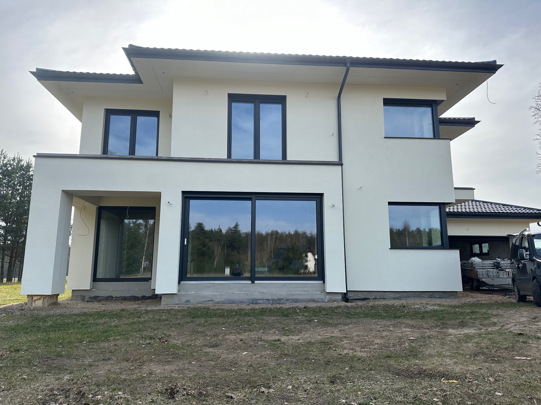 Nowoczesny, dwukondygnacyjny dom z białymi ścianami i czarnymi ramami okiennymi. Widoczne elementy wykończeniowe elewacji i teren wokół domu w trakcie zagospodarowania.