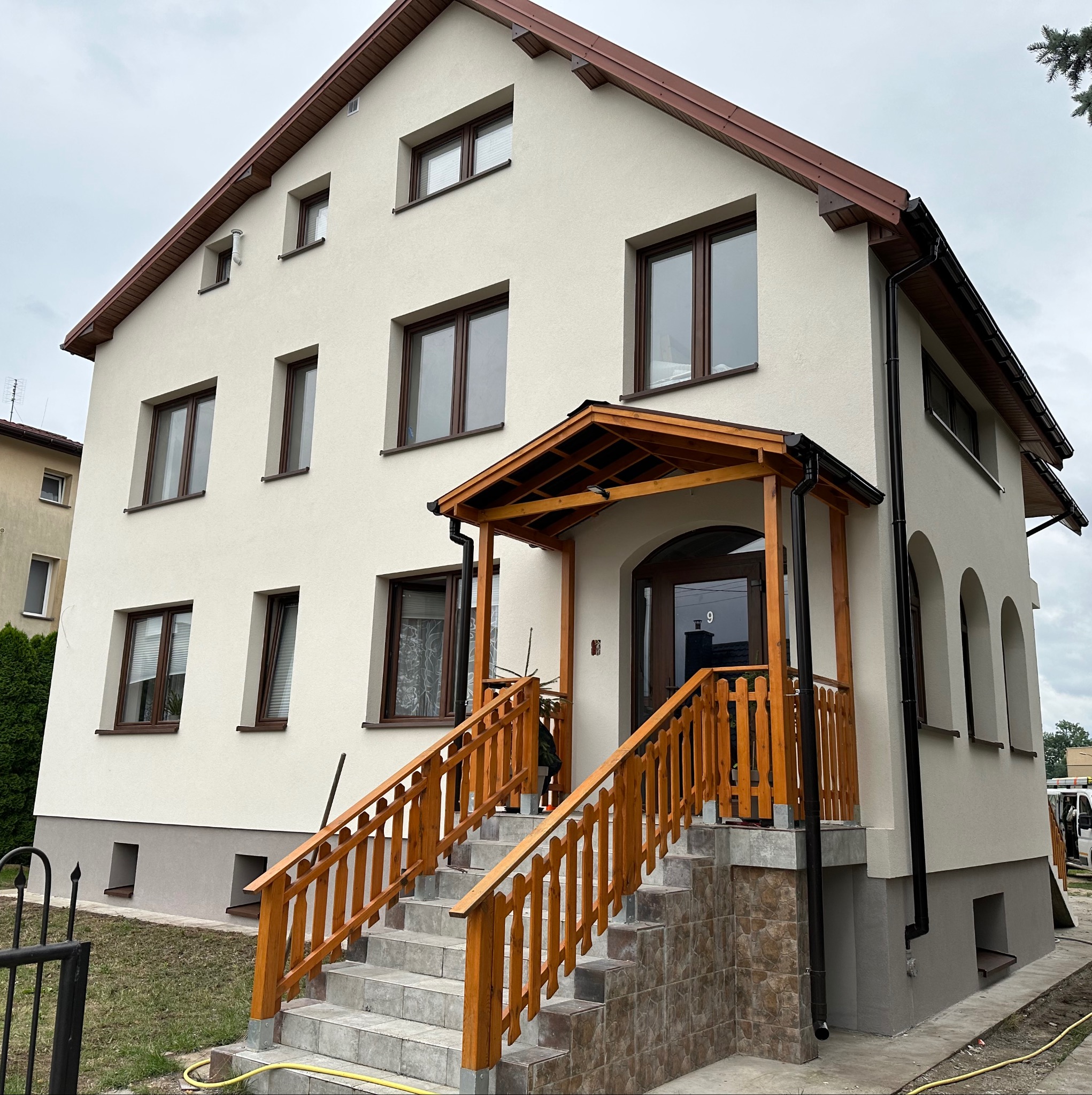 Dwupiętrowy dom z jasną elewacją i brązowymi oknami, drewnianym gankiem i schodami. Widoczne rynny i podmurówka w szarym kolorze. Pochmurne niebo w tle.