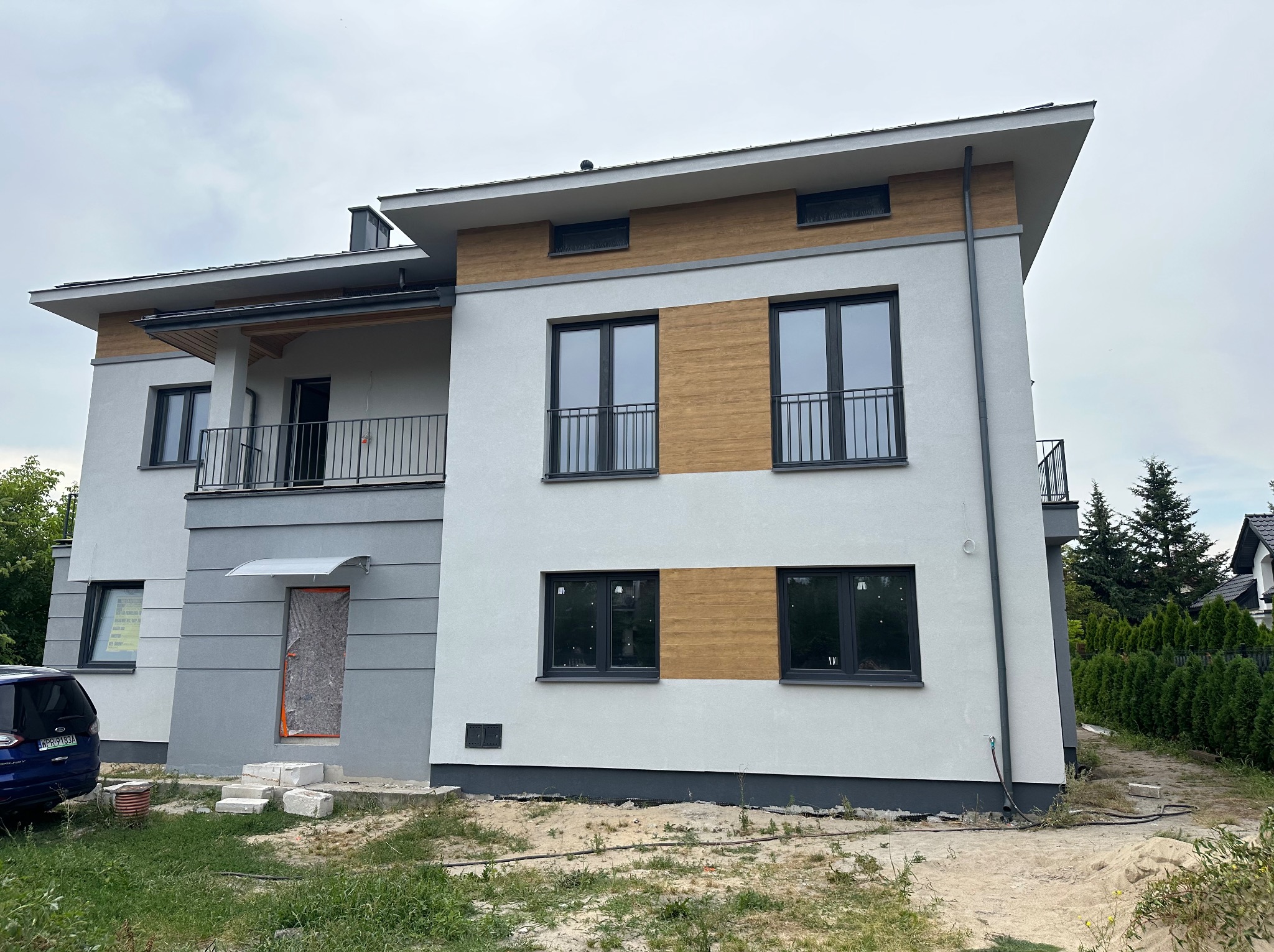 Nowoczesny dom z jasną elewacją, drewnianymi akcentami i czarnymi oknami. Część elewacji w szarym kolorze. Widoczne elementy wykończeniowe i teren wokół budynku.