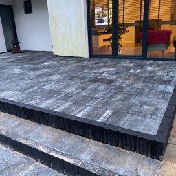 Nik-bruk - Nowoczesny taras z kostki w odcieniach szarości, obrzeża z ciemnej kostki, widoczne trzy stopnie, w tle przeszklone drzwi tarasowe z widokiem na wnętrze domu.