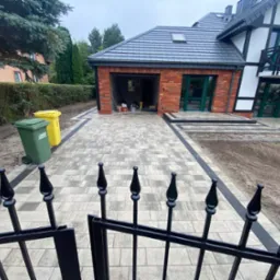 Ułożona kostka brukowa w odcieniach szarości z czarnymi obrzeżami prowadząca do garażu z cegły i domu z balkonem, widok zza czarnego, metalowego ogrodzenia.