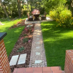 Ogród z ścieżką z kostki granitowej prowadzącą do drewnianego stołu z ławkami, otoczony zielenią i rabatami kwiatowymi, widok z perspektywy schodów.