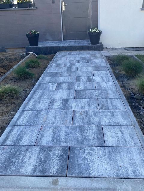 Ścieżka z prostokątnej kostki brukowej w odcieniach szarości prowadząca do drzwi wejściowych, ozdobiona donicami z kwiatami po bokach, z widocznymi elementami trawnika w trakcie zakładania.