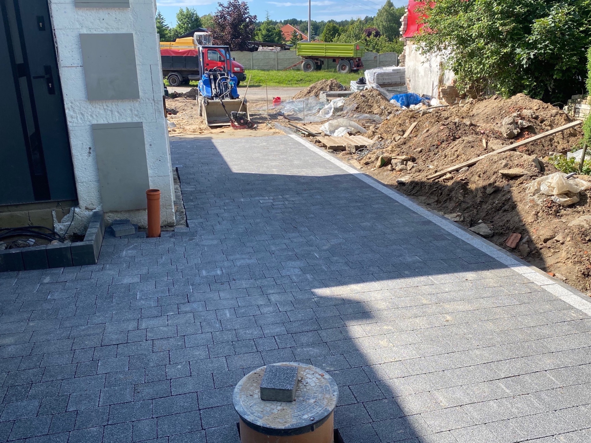 Ułożona szara kostka brukowa z białym obrzeżem, widoczny mini-ładowarka i wywrotka w tle, teren budowy w trakcie prac ziemnych.