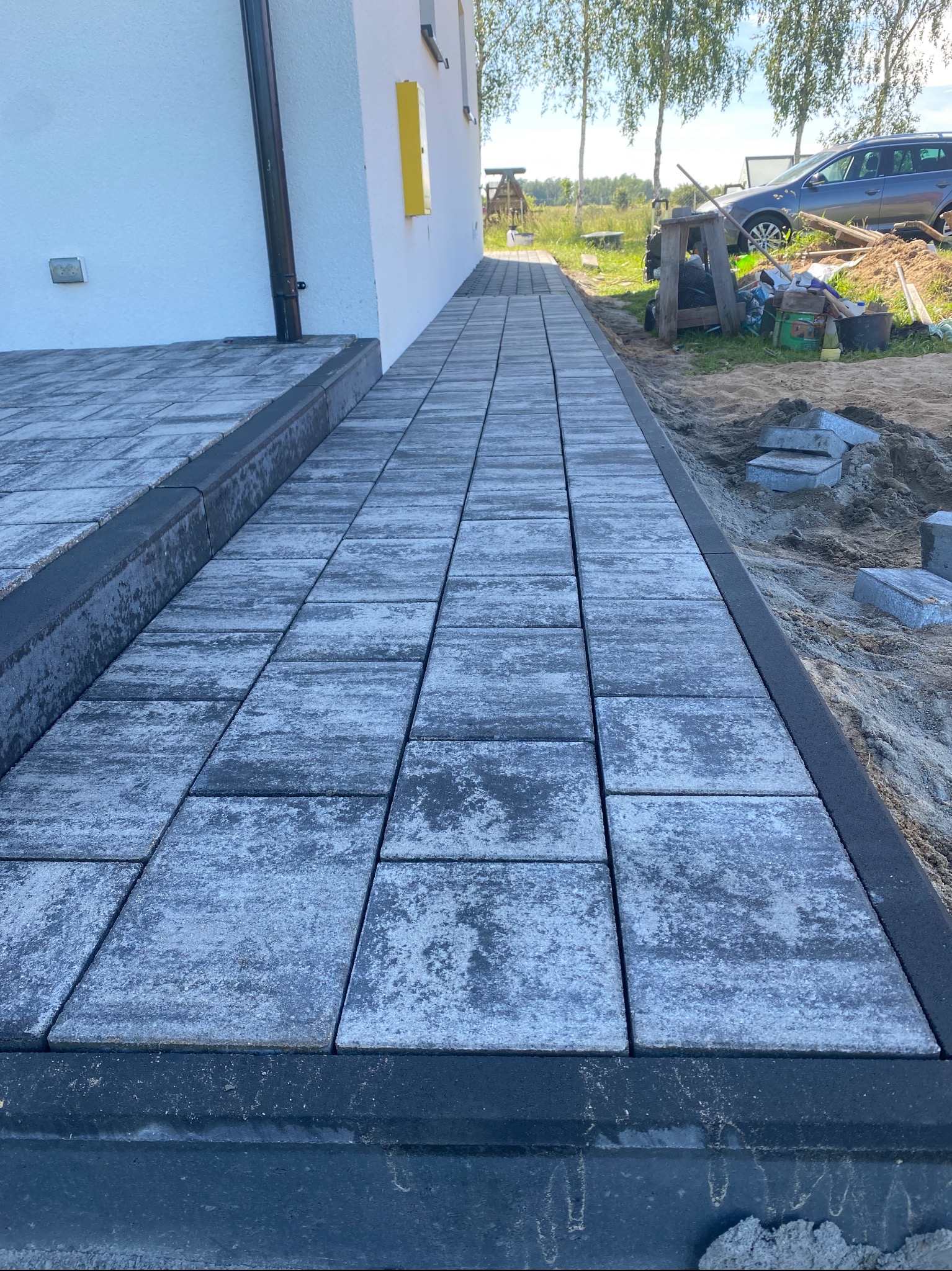 Świeżo ułożony chodnik z szarej kostki brukowej, obrzeża z ciemnego betonu, w tle dom z żółtym akcentem i rozkopany teren budowy.