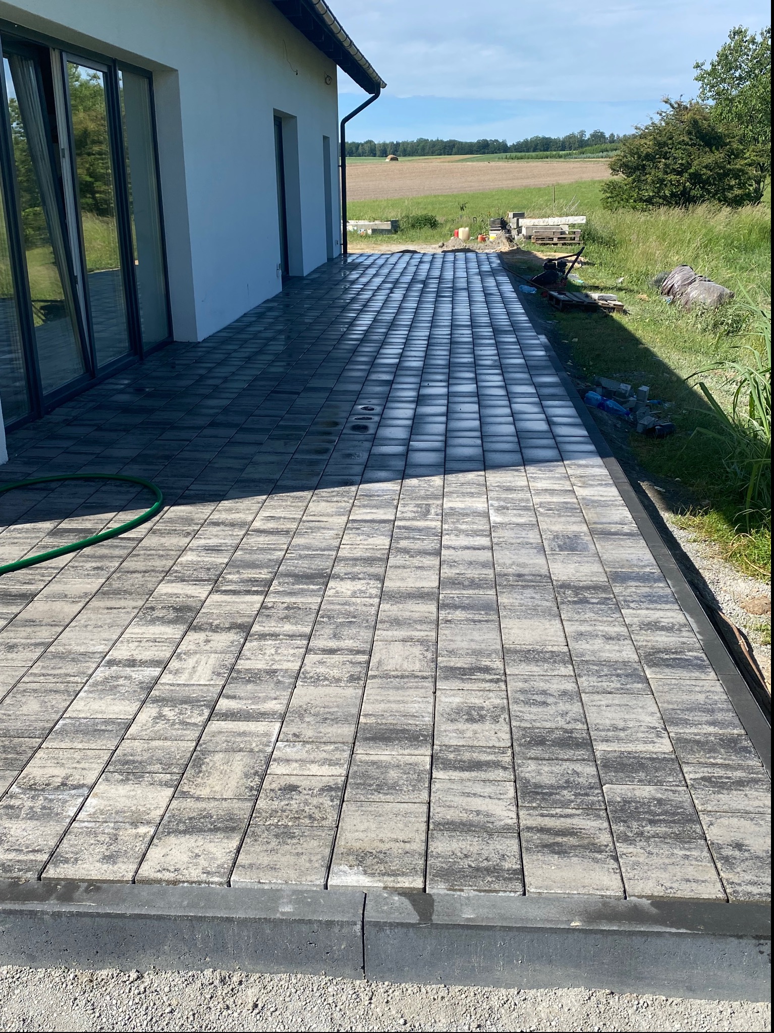 Wyłożony kostką taras przy nowoczesnym domu z dużymi oknami przesuwnymi, widok na pola w tle, częściowo ocieniony, z zielonym wężem ogrodowym na kostce.