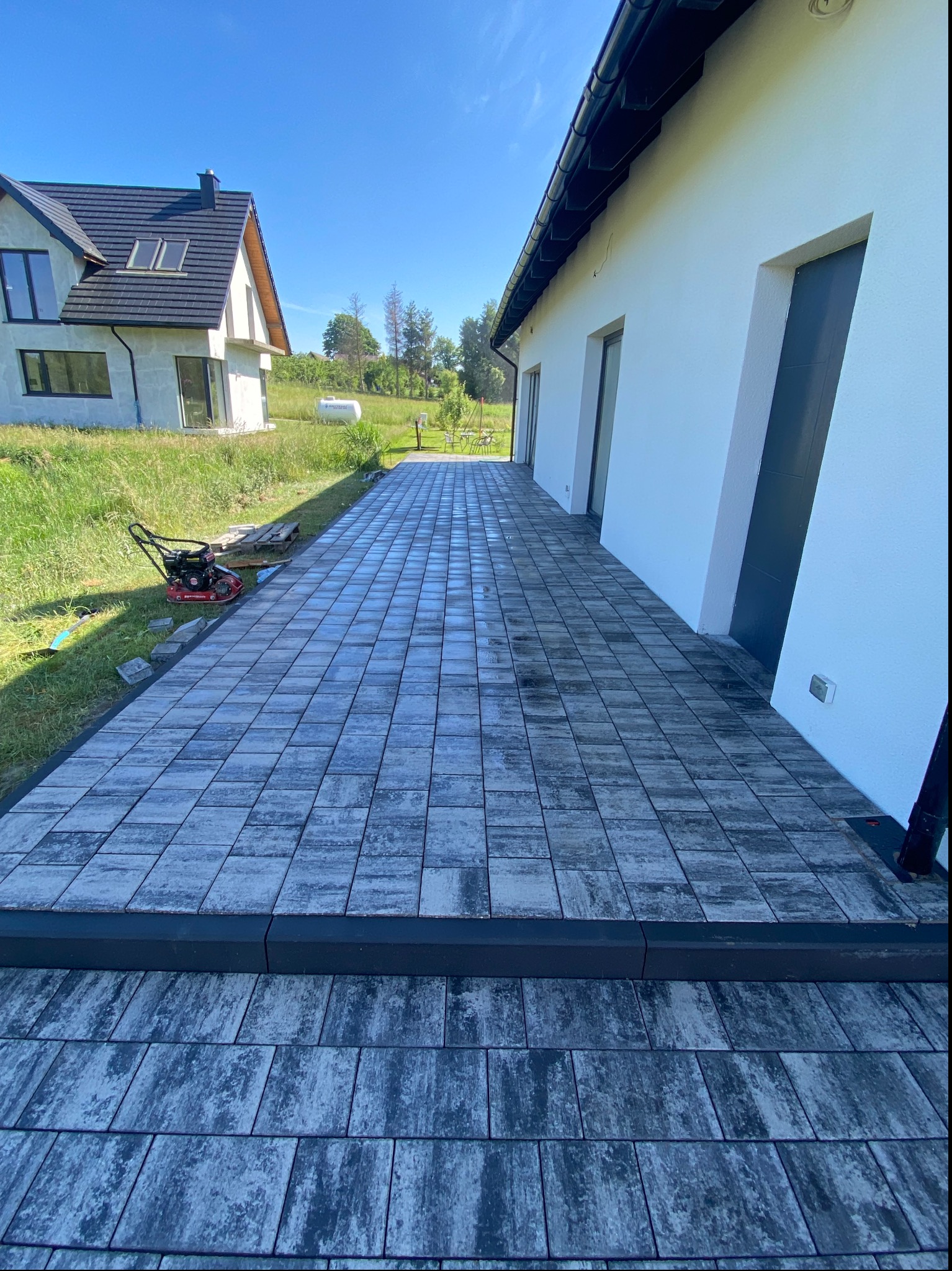 Ułożona szara kostka brukowa tworząca ścieżkę wzdłuż białego budynku z ciemnymi drzwiami, w tle dom z ciemnym dachem i ubijarka wibracyjna.