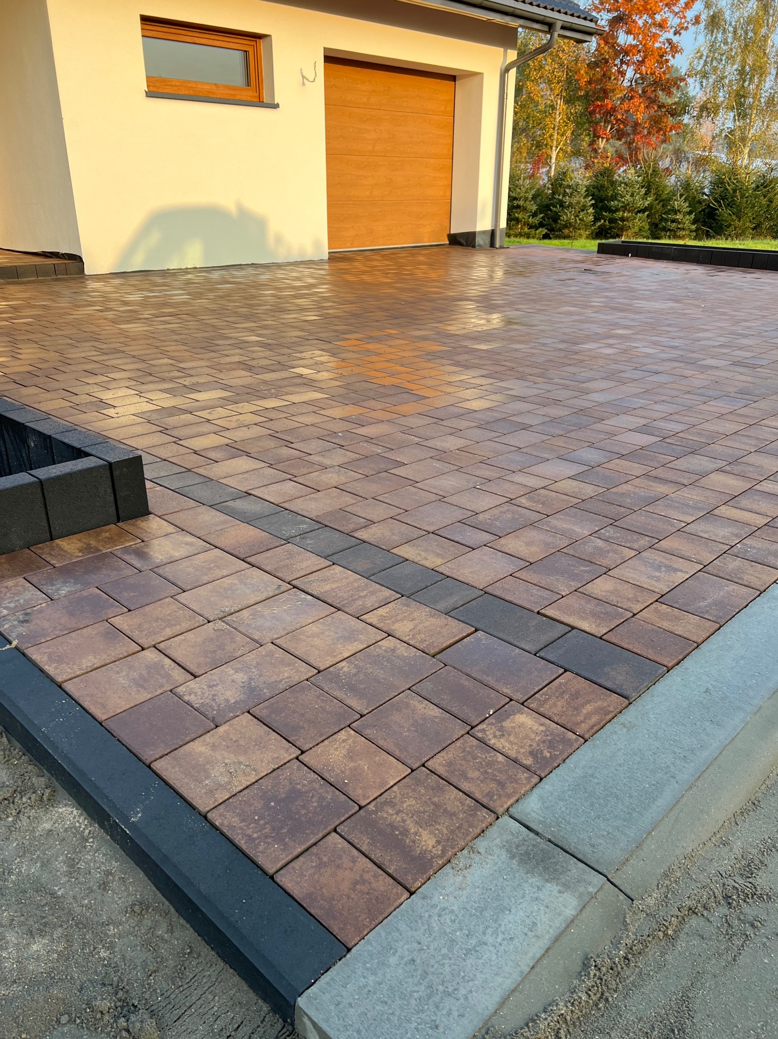Nowa nawierzchnia z kostki brukowej w odcieniach brązu i szarości, obrzeża z kostki betonowej, widok na garaż i fragment budynku w tle, jesienne drzewa w oddali.