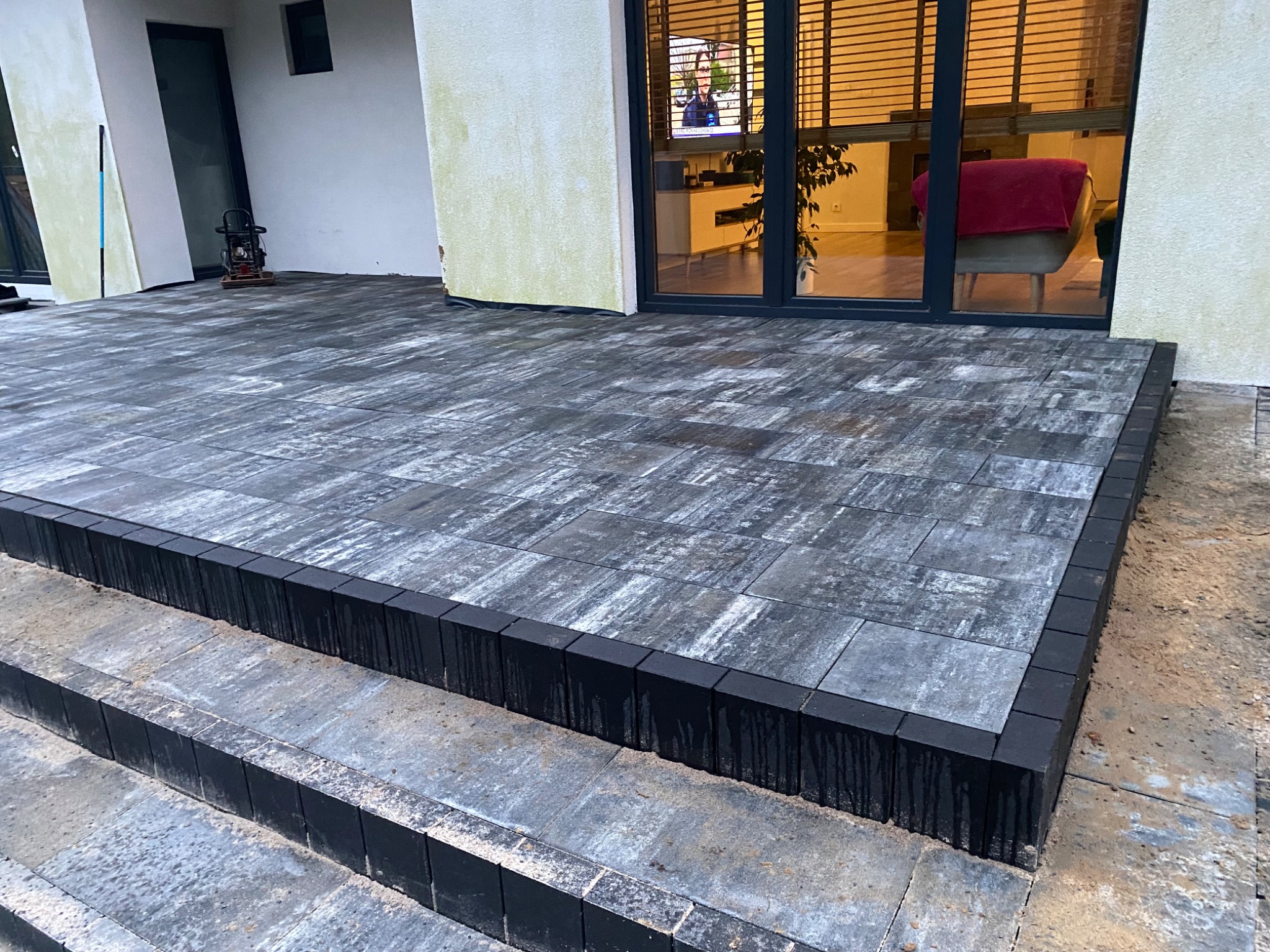 Nowoczesny taras z kostki w odcieniach szarości, obrzeża z ciemnej kostki, widoczne trzy stopnie, w tle przeszklone drzwi tarasowe z widokiem na wnętrze domu.