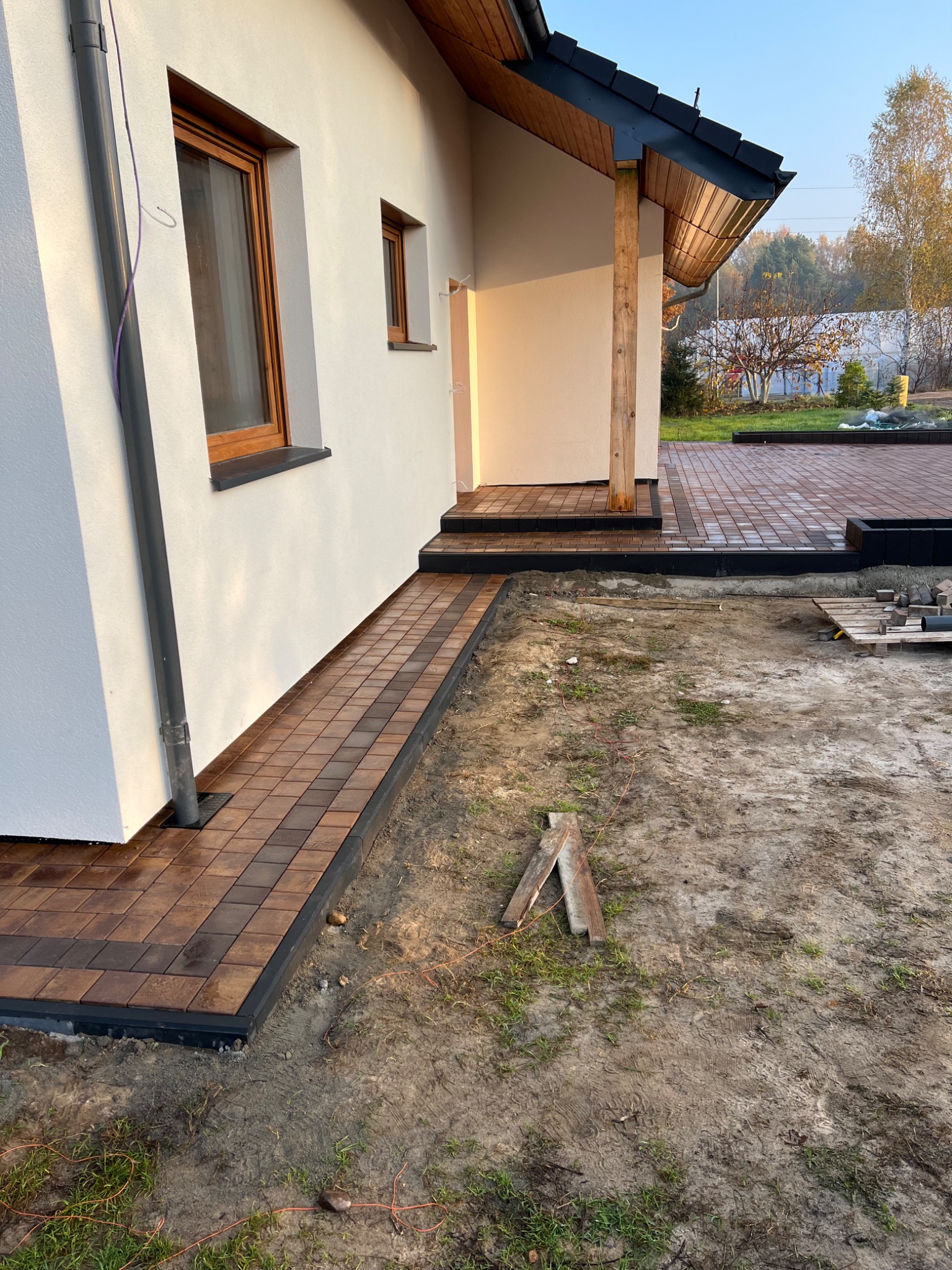 Ułożona ścieżka z kostki brukowej prowadząca wzdłuż białej ściany domu, widoczne przygotowane podłoże i fragment tarasu z kostki w tle.